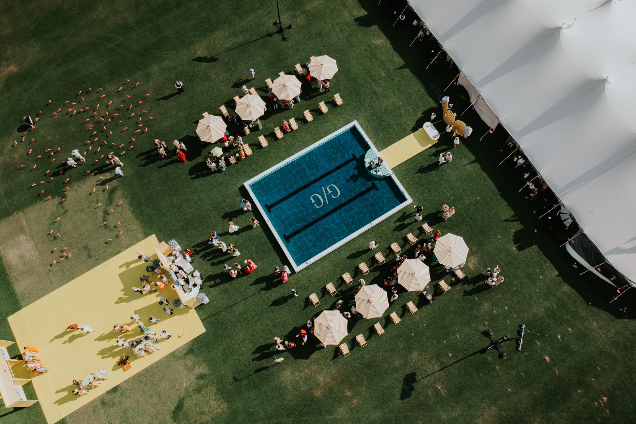 Aerial view of an outdoor event with a pool, yellow pathways, striped umbrellas, and people gathered on a grassy area. Large white tent visible on the right.