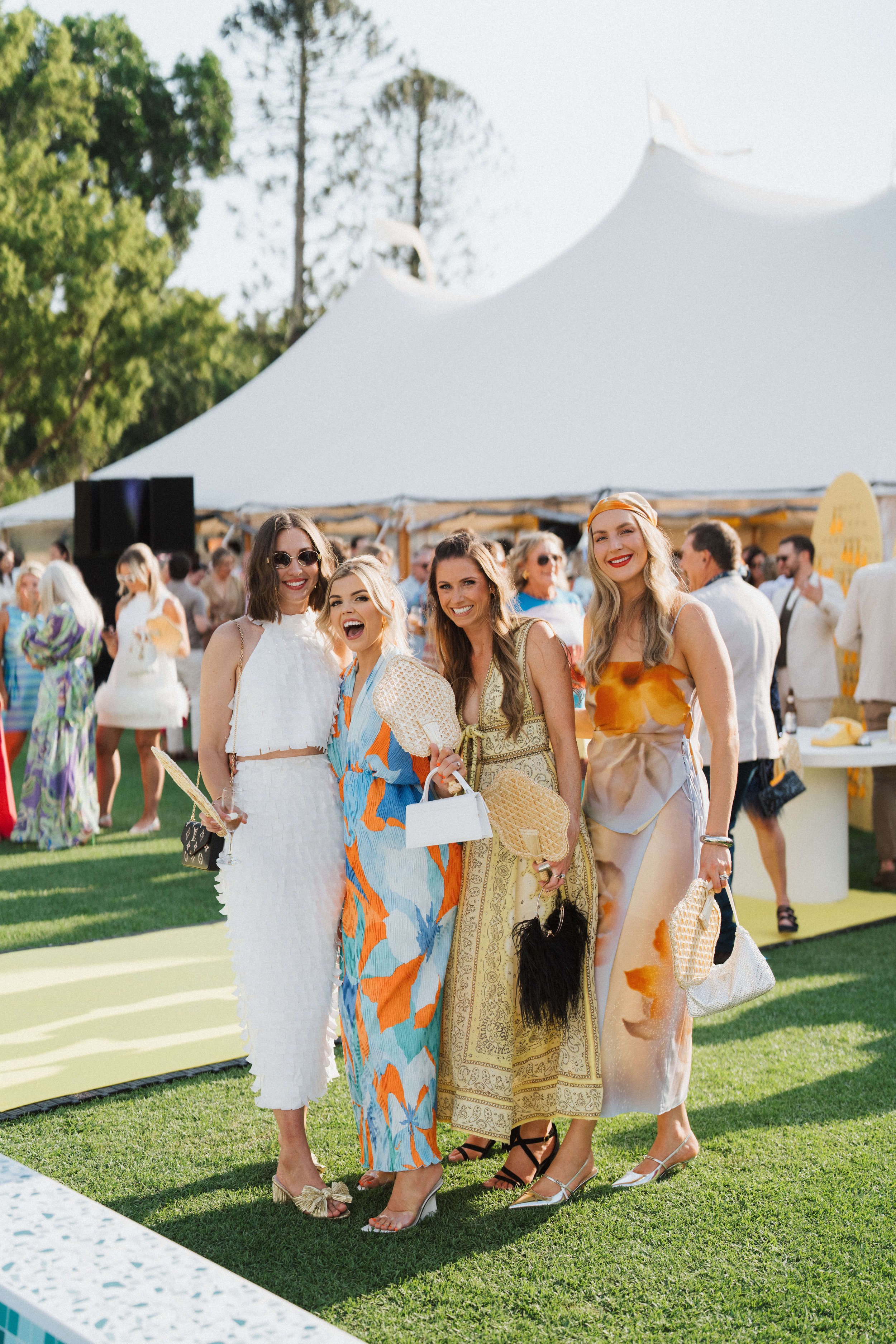 Group of women in colorful dresses smiling outdoors at a social event with a tent and crowd in the background.