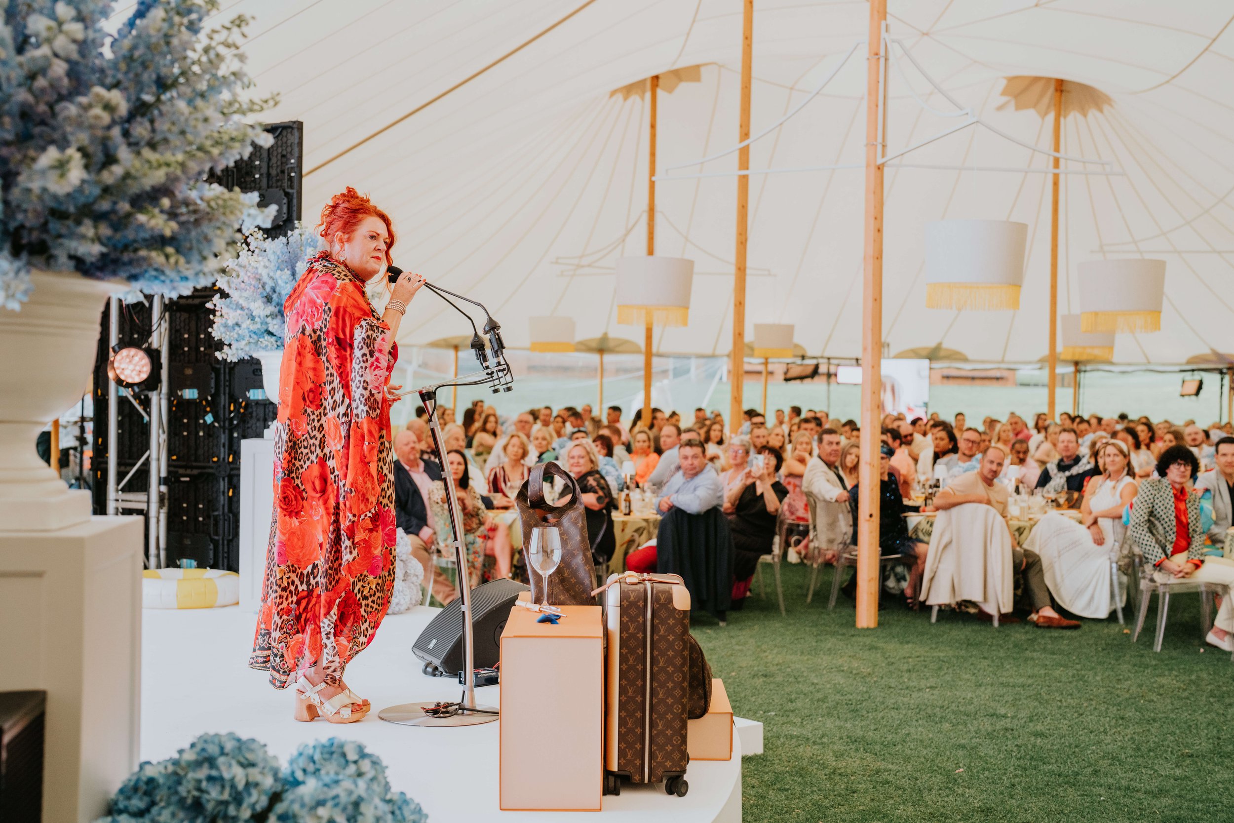 A person speaking on stage at an event with an attentive audience seated in a tent. The speaker wears a colorful patterned outfit, and luggage is placed on stage. The tent is decorated with large flower arrangements and pendant lights.