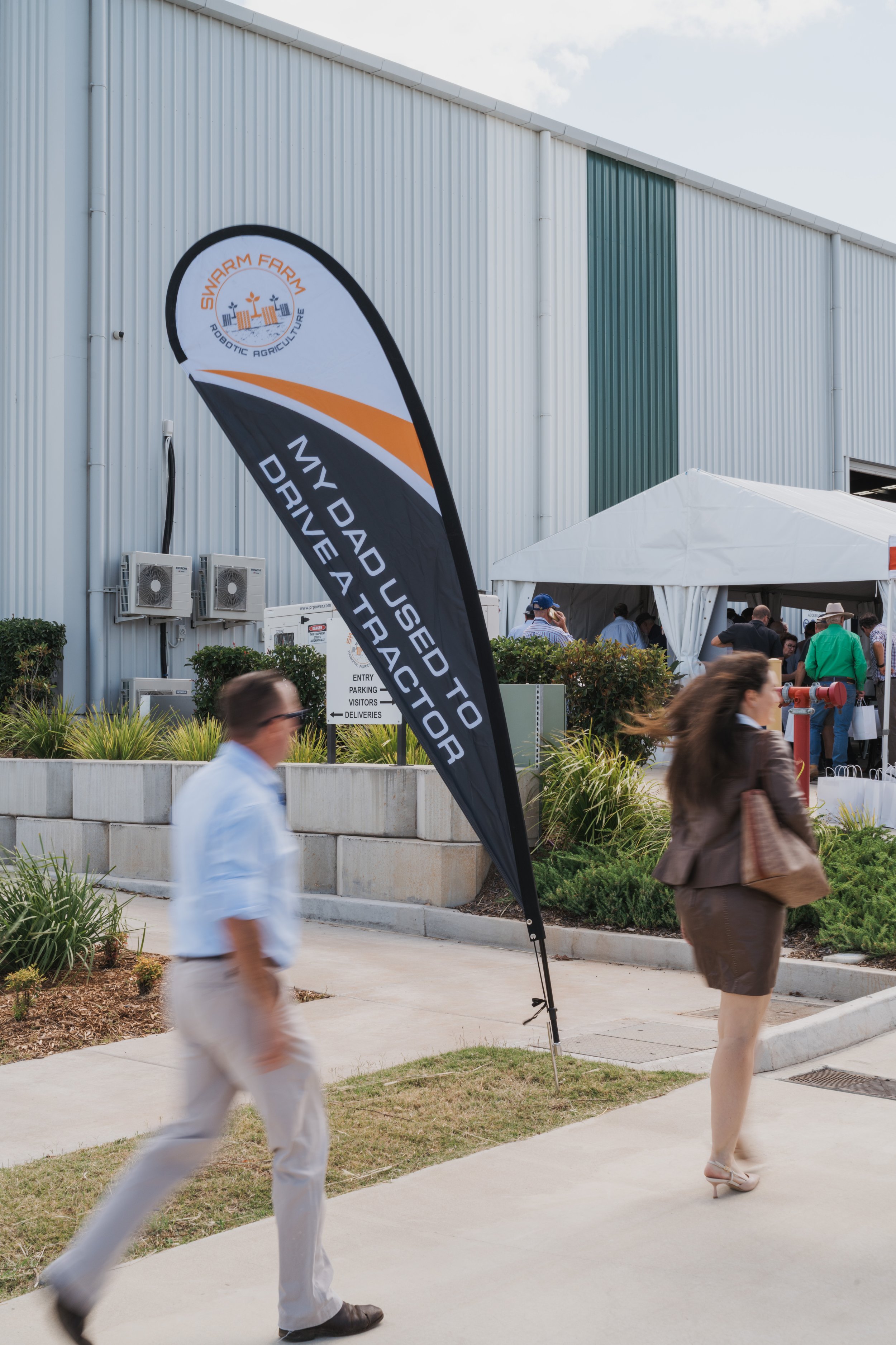 A banner at an event outside a building reads 'My dad used to drive a tractor' with the logo 'Swarm Farm Robotic Agriculture.' Two people walk past, and tents and attendees are visible in the background.