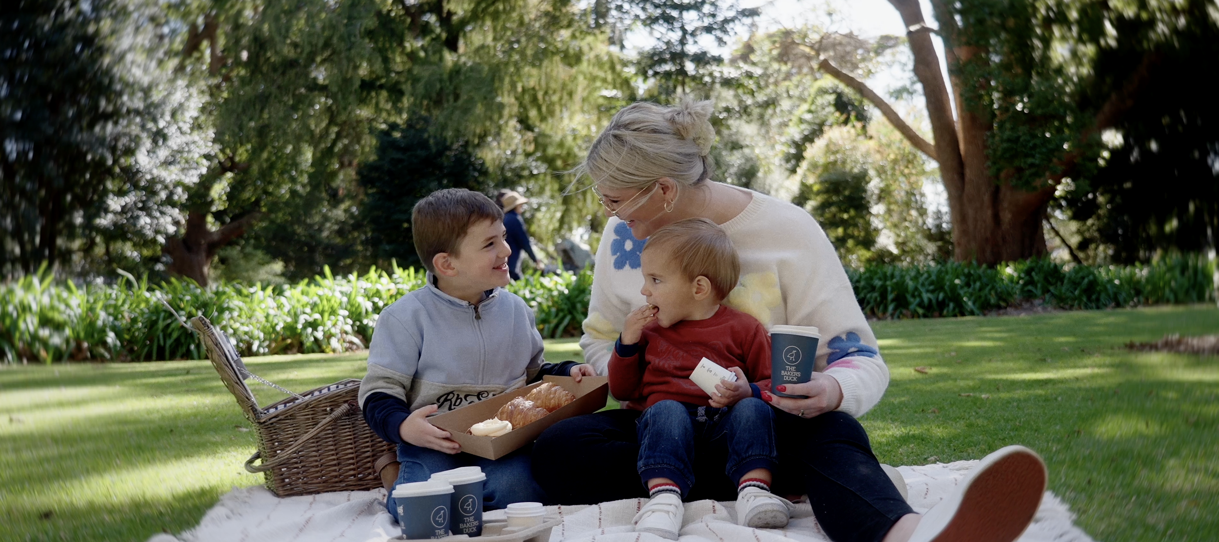 Family picnic in a park with a mother and two children on a blanket, smiling and enjoying pastries and drinks, surrounded by greenery and trees.