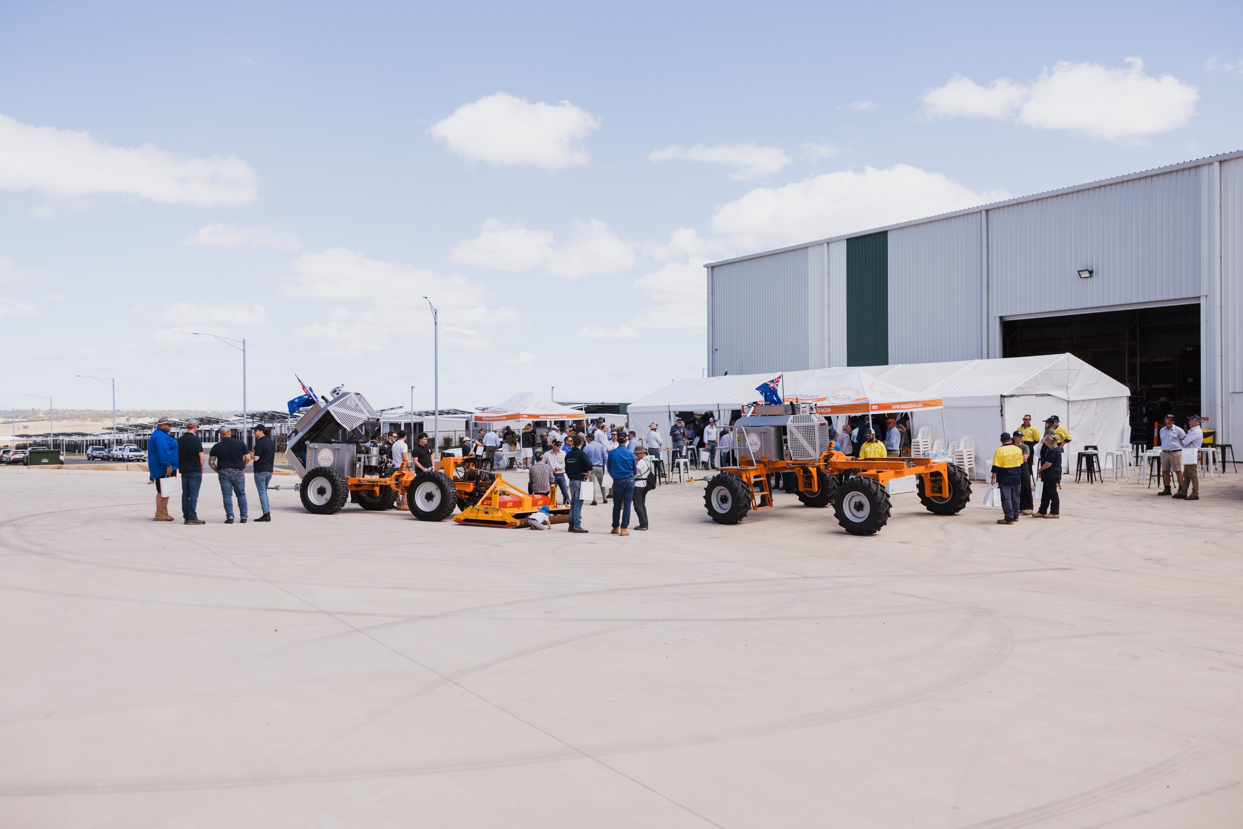 People gathered around agricultural machinery outside large tents and a warehouse on a sunny day.