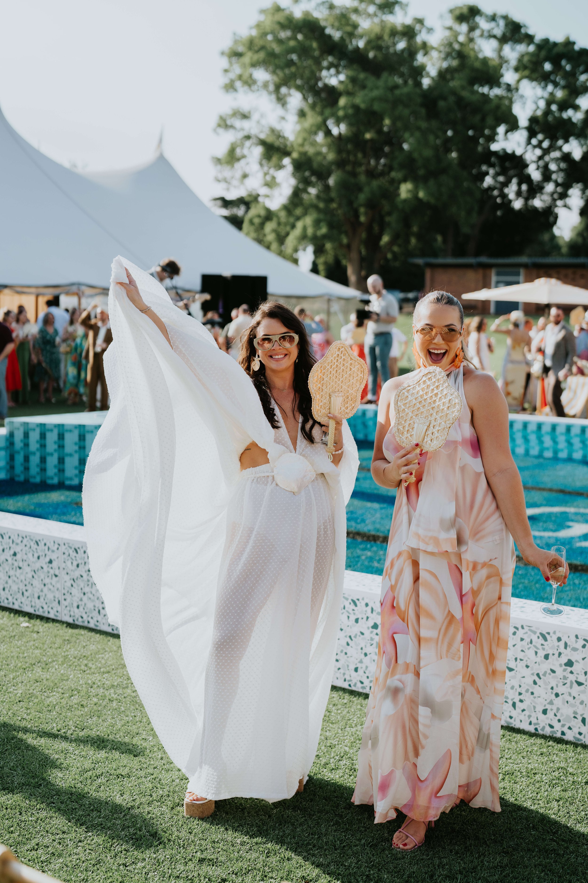 Two women in elegant summer dresses smiling near a pool at an outdoor event, holding hand fans. In the background, attendees gather under a large white tent.