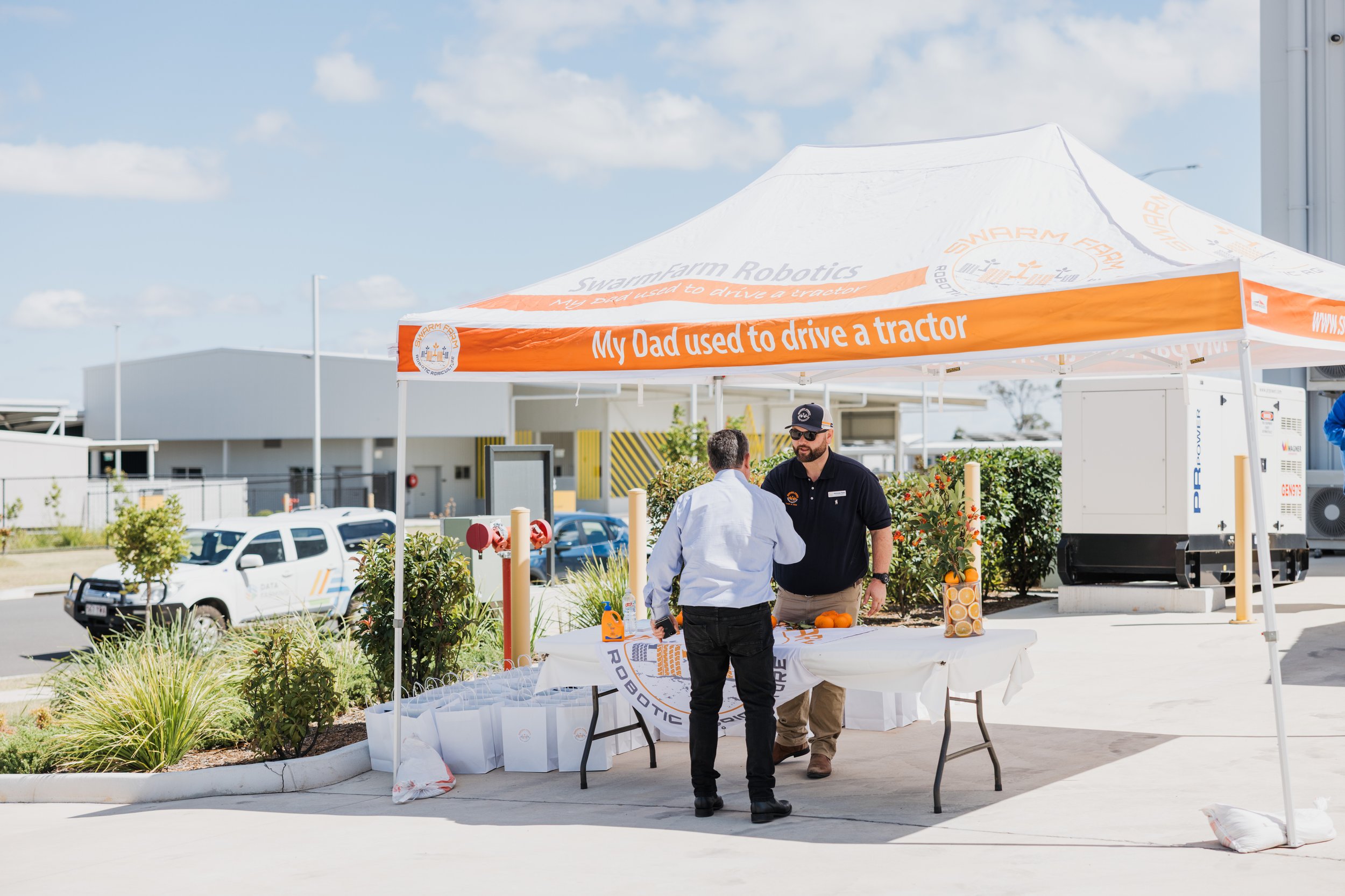 Two people standing under a white and orange tent labeled 'Swarmfarm Robotics' with 'My Dad used to drive a tractor.' Table with items and promotional bags visible.