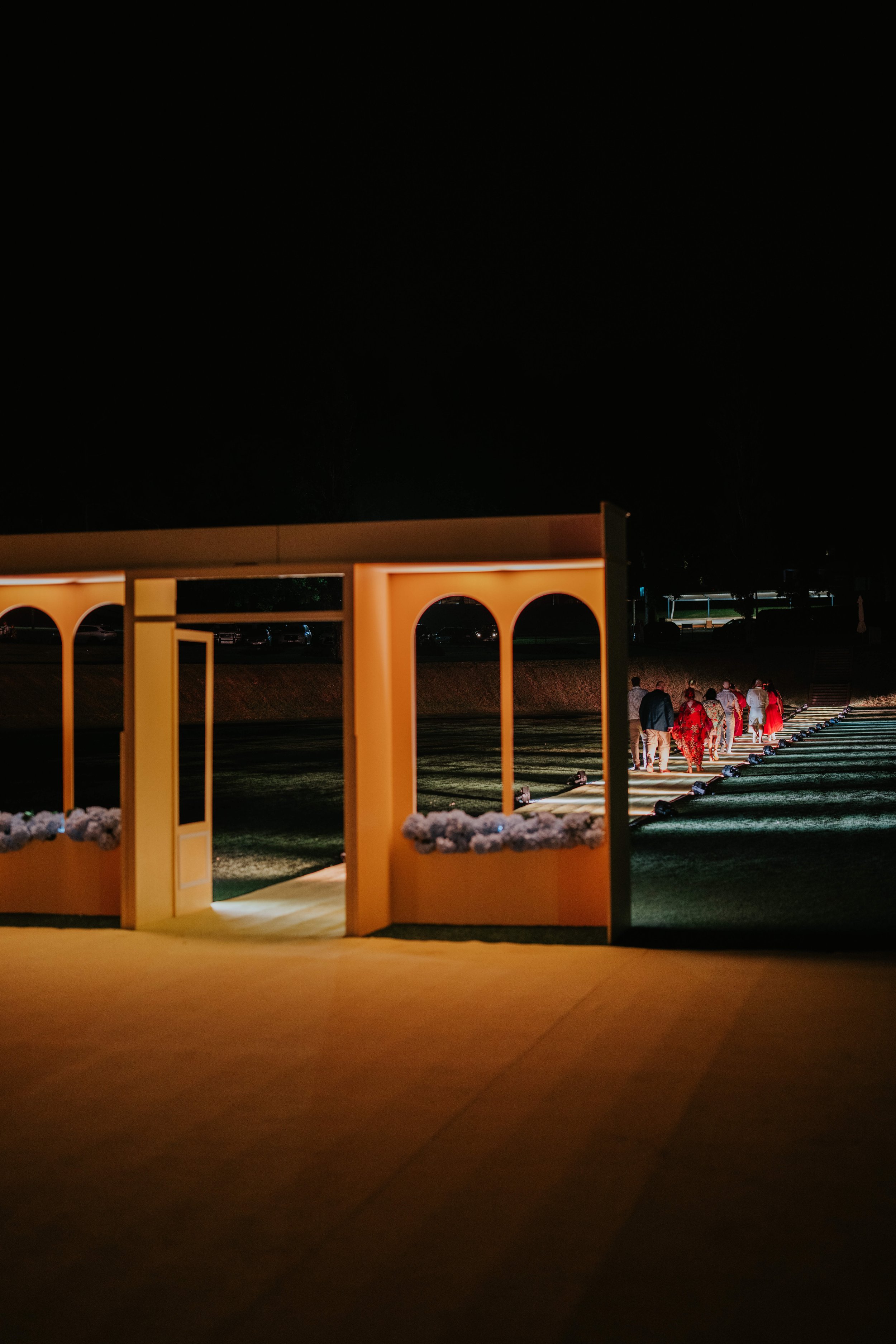 Illuminated walkway with open structure, floral decorations, and a group of people walking on a path at night.