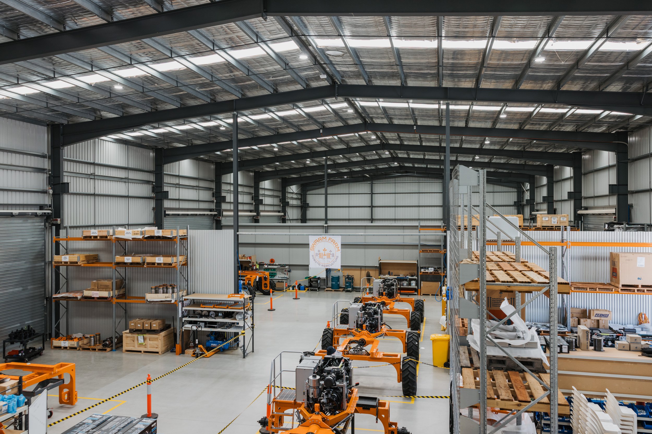 Interior of a large warehouse with industrial equipment and shelves. Various orange machinery and stacked boxes are visible, along with metal shelving and a loft area. The space is organized with visible aisles and equipment on the floor.