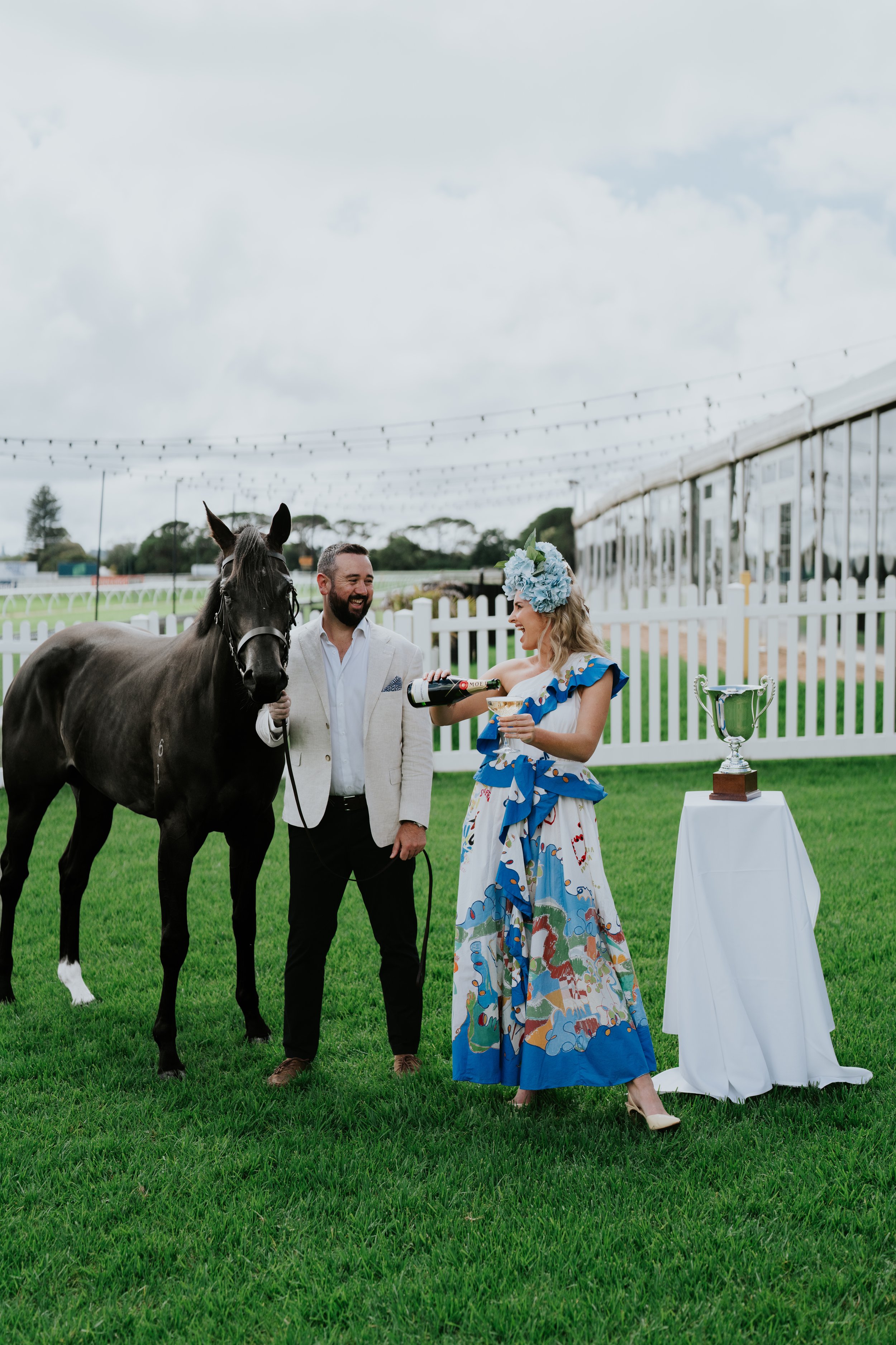 A man and a woman standing beside a horse on a grassy field, with the woman pouring champagne into a glass. A trophy sits on a table with a white cloth nearby, and string lights are overhead against a cloudy sky.