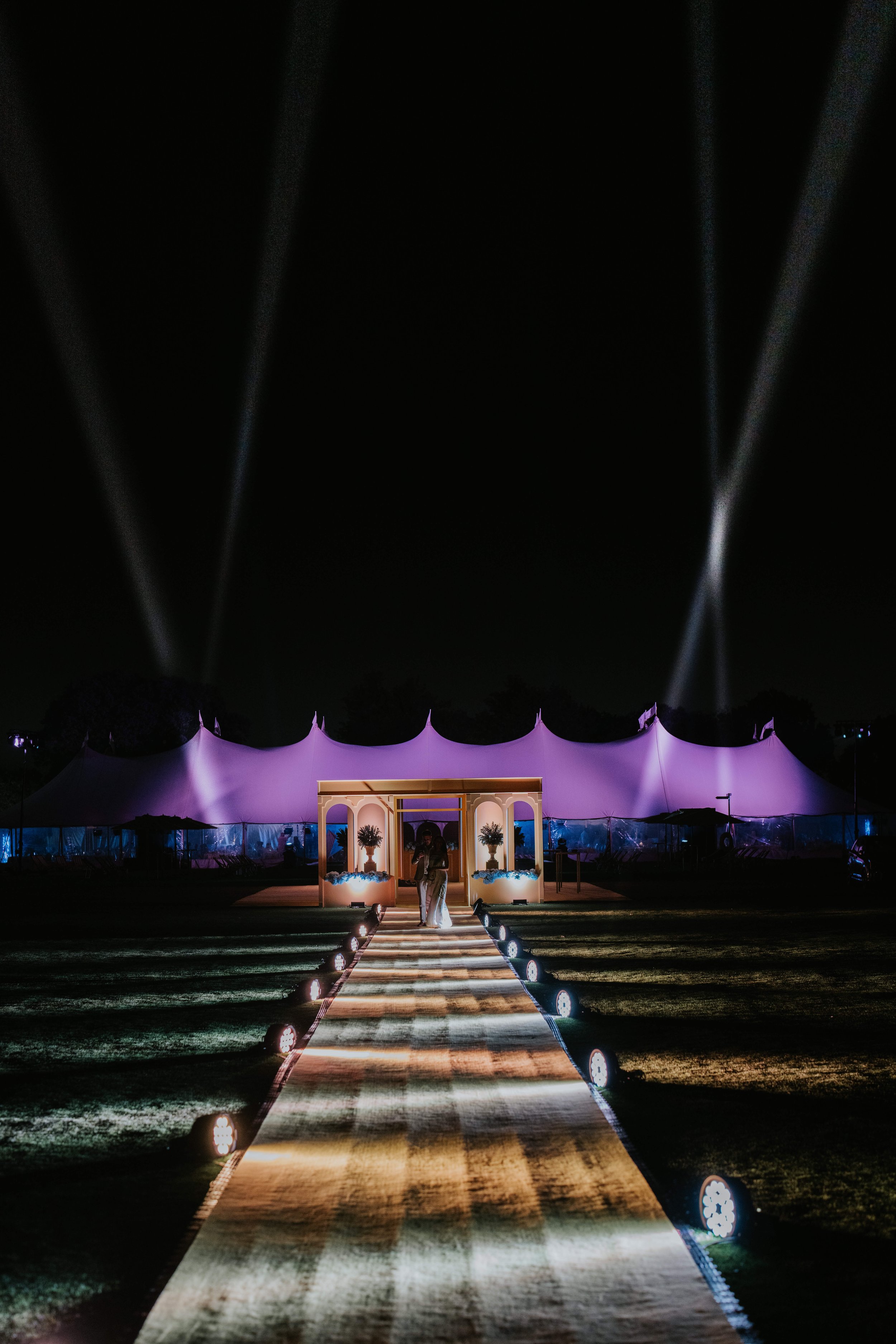 Nighttime event with illuminated pathway leading to a large tent, spotlight beams in the sky, and decorative entrance with plants.