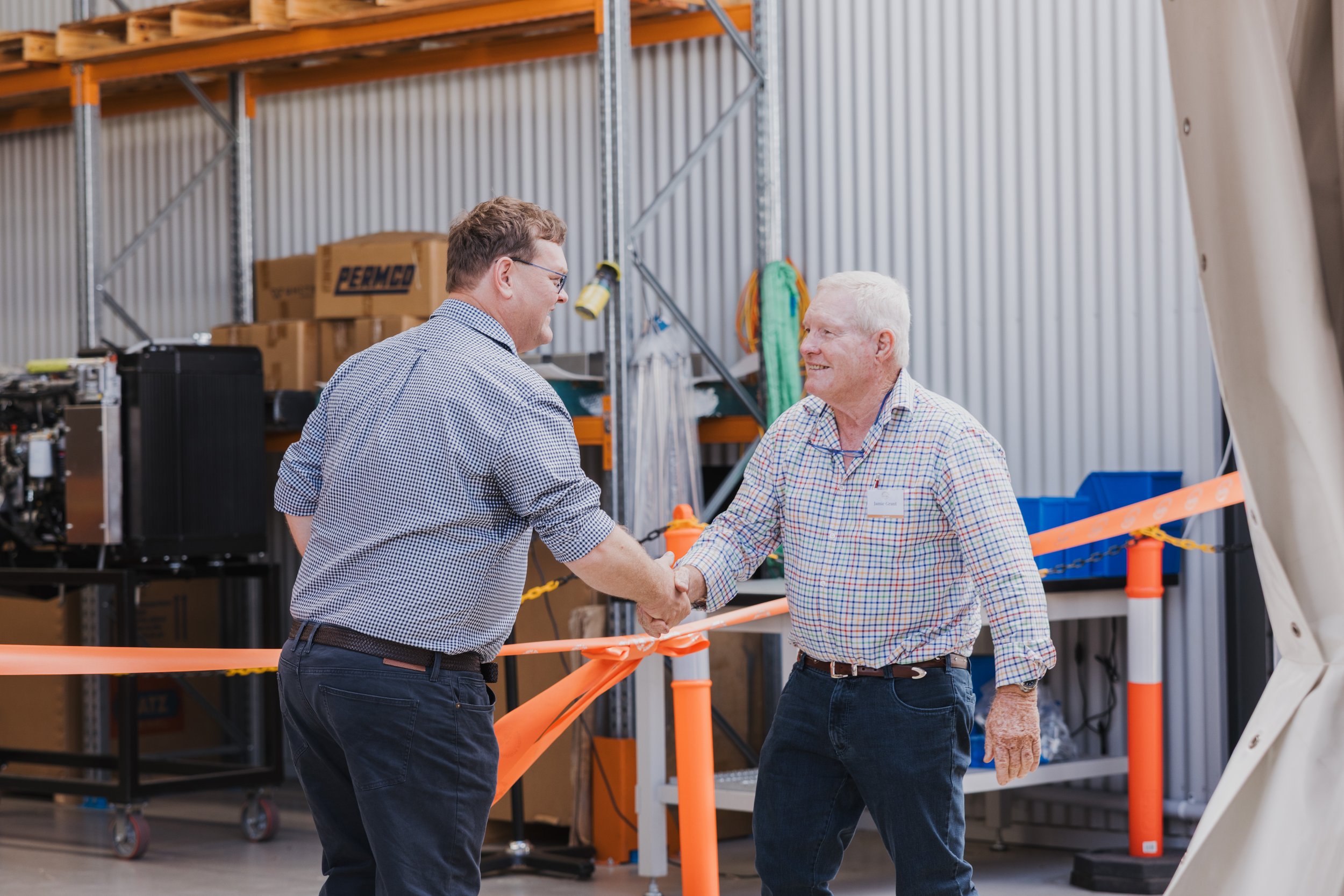 Two men shaking hands at an industrial facility with boxes and equipment in the background.
