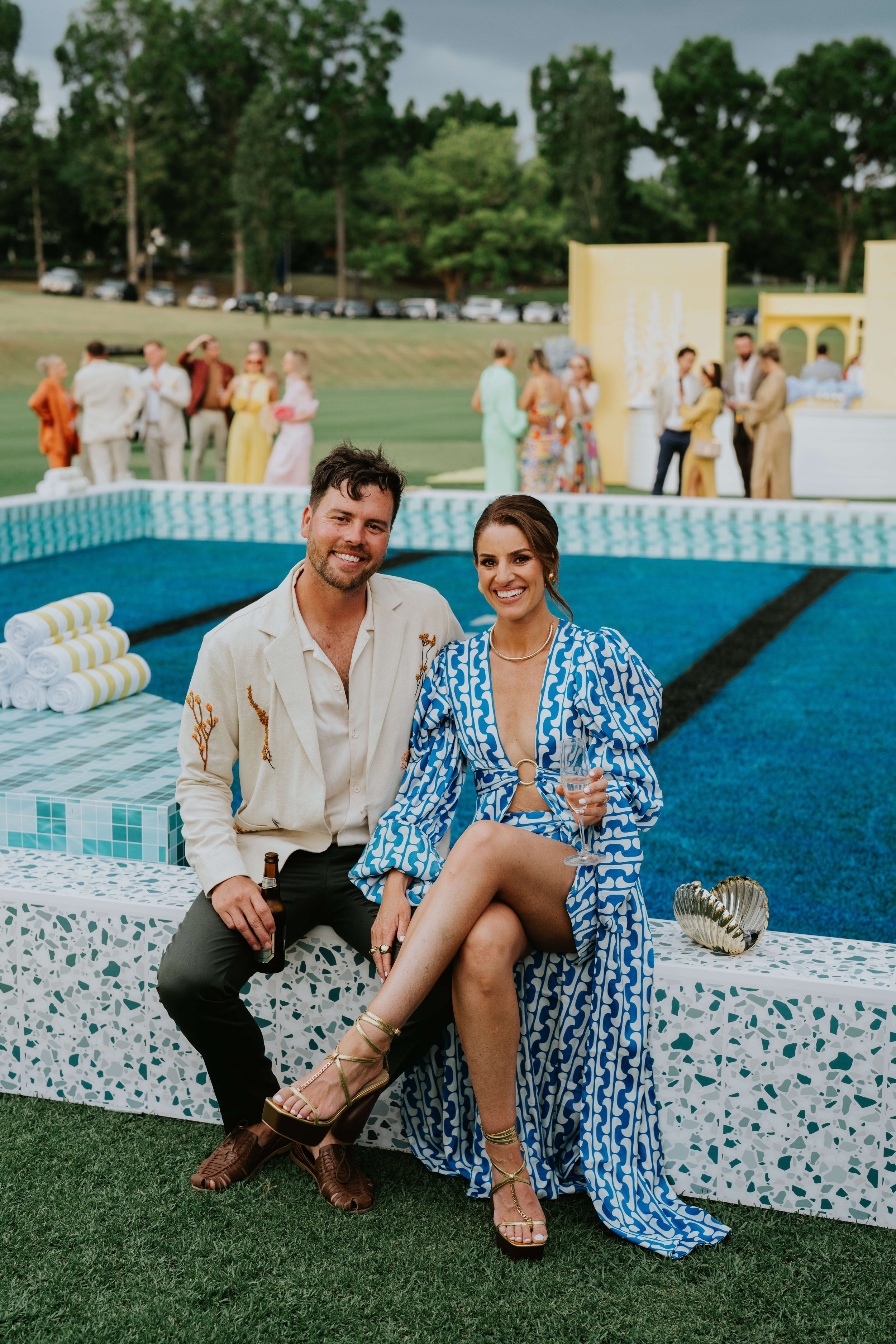 A man and woman sitting by a pool, smiling, with people socializing in the background. The woman is wearing a blue patterned dress and holding a drink, while the man is wearing a light-colored shirt and holding a beer.