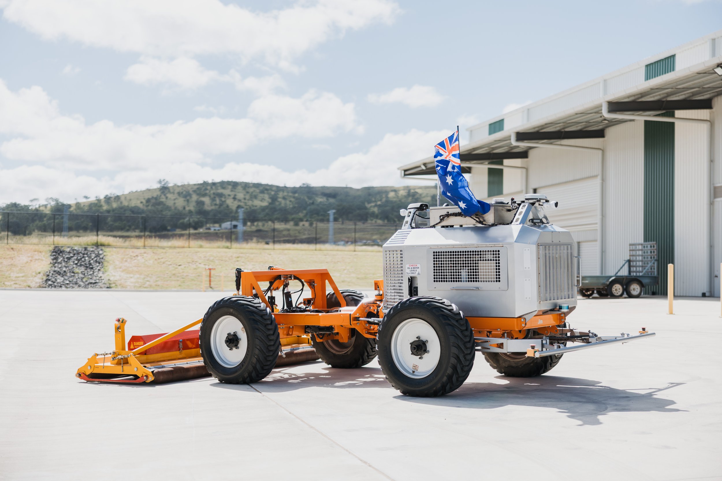 Autonomous agricultural vehicle on concrete yard with an attached implement, flying an Australian flag, in front of industrial buildings and a rural landscape.