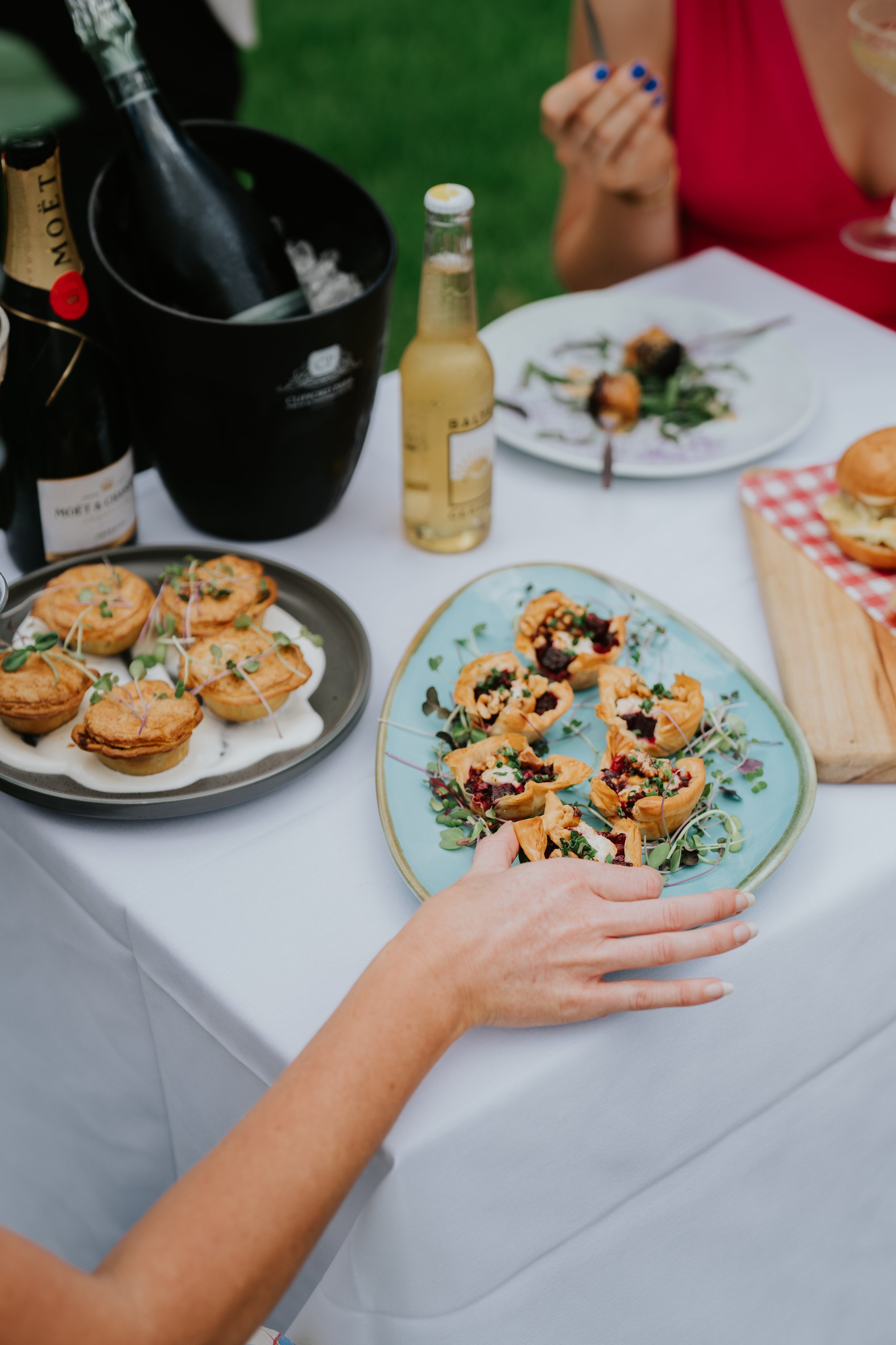 An outdoor table setting with plates of appetizers, including pastries and small tarts, alongside a bottle of champagne in an ice bucket, a beer bottle, and a plate with a small salad. A person's hand is reaching for food.