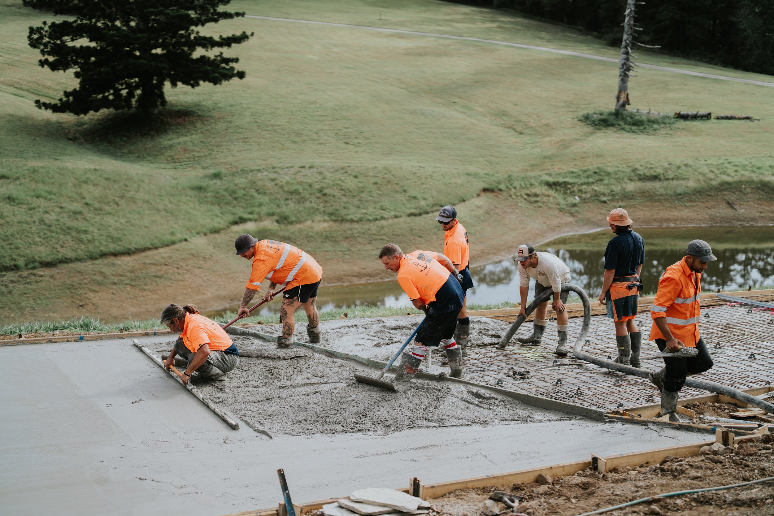 Construction workers wearing orange safety vests pouring and smoothing concrete on a large outdoor slab, in a grassy area with trees in the background.