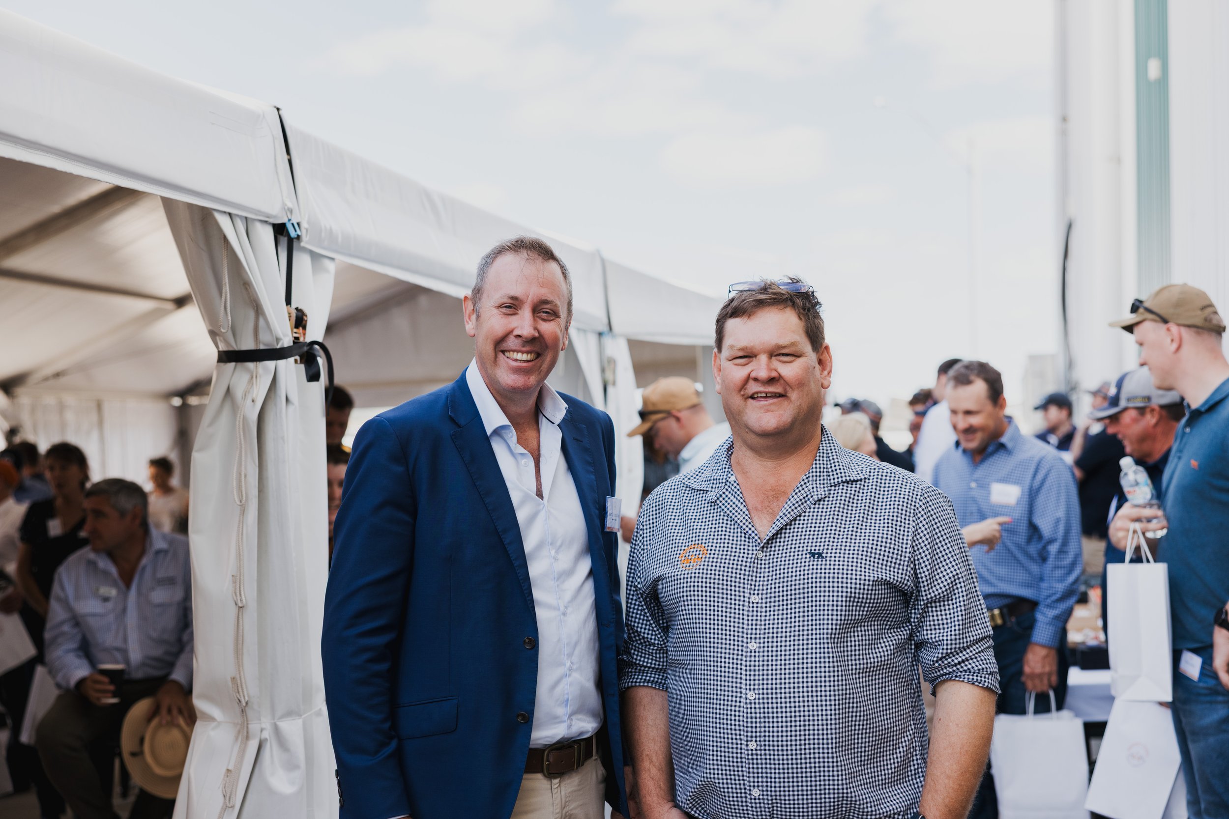 Two men smiling at an outdoor event under a tent, surrounded by other people in casual attire.