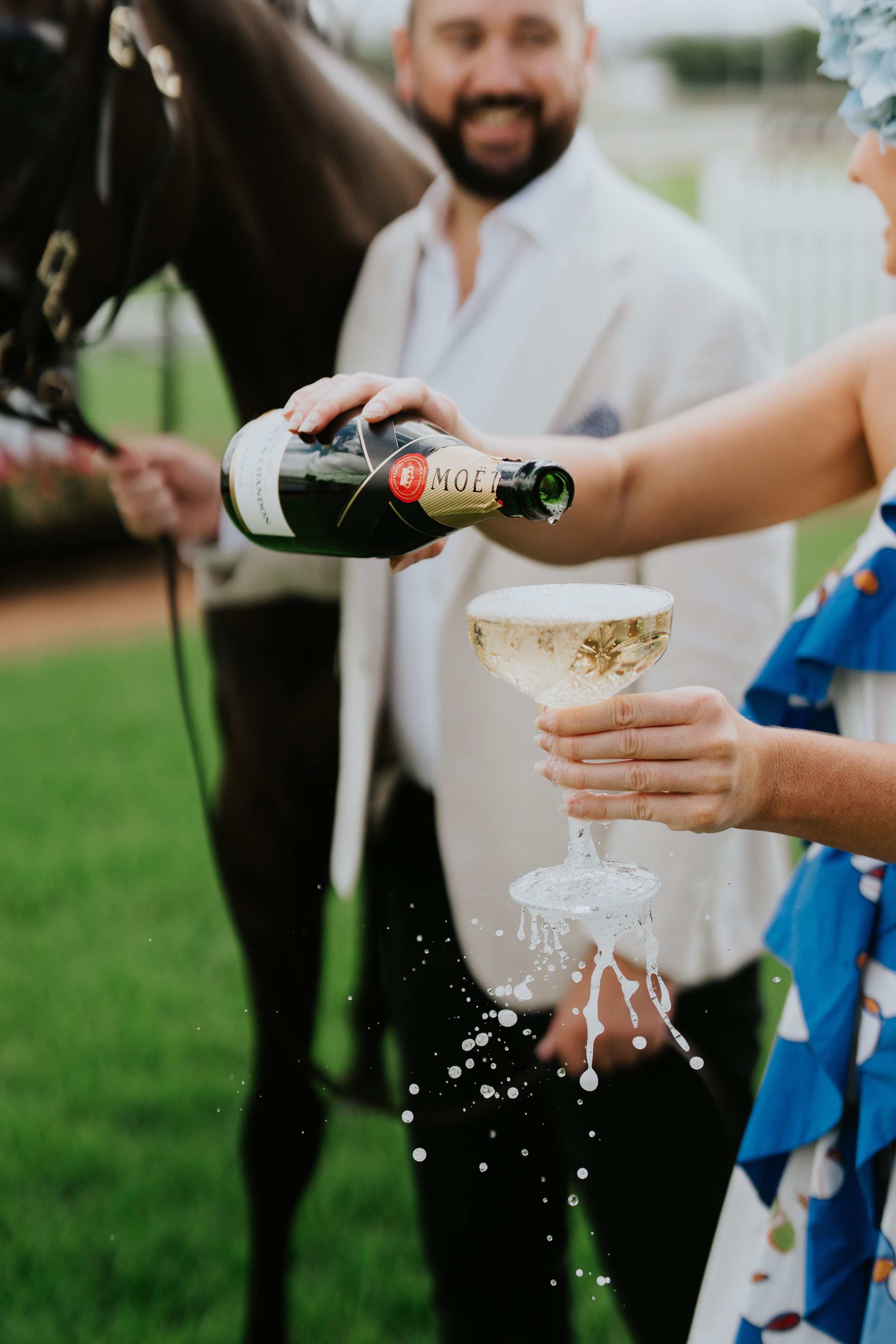 Person pouring champagne into a glass, outdoors, with a horse and a smiling individual in the background.