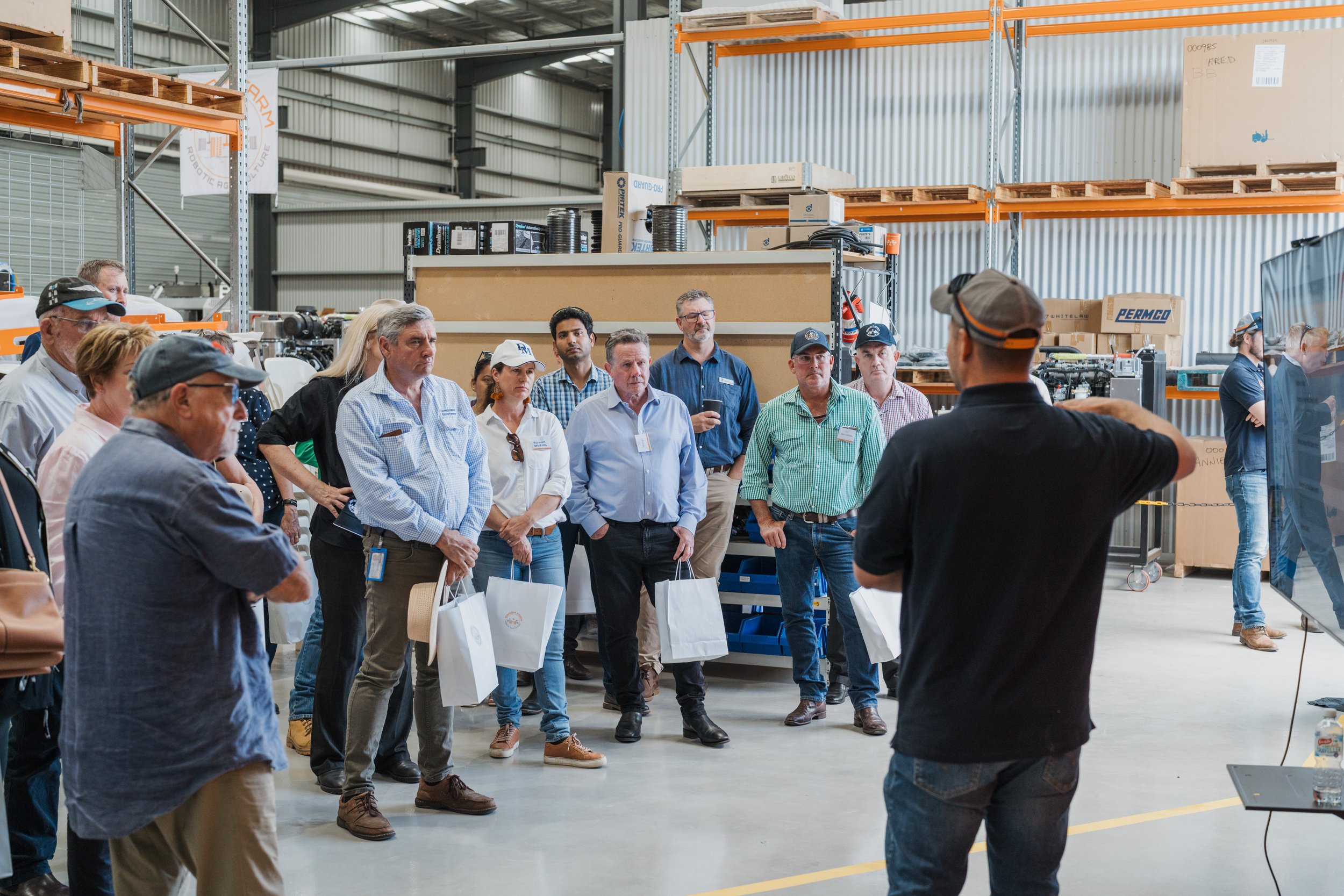 A group of people in a warehouse setting attending a presentation, with an instructor pointing to a screen or machinery. They are dressed in casual business attire, holding gift bags. Shelving with boxes and equipment is visible in the background.