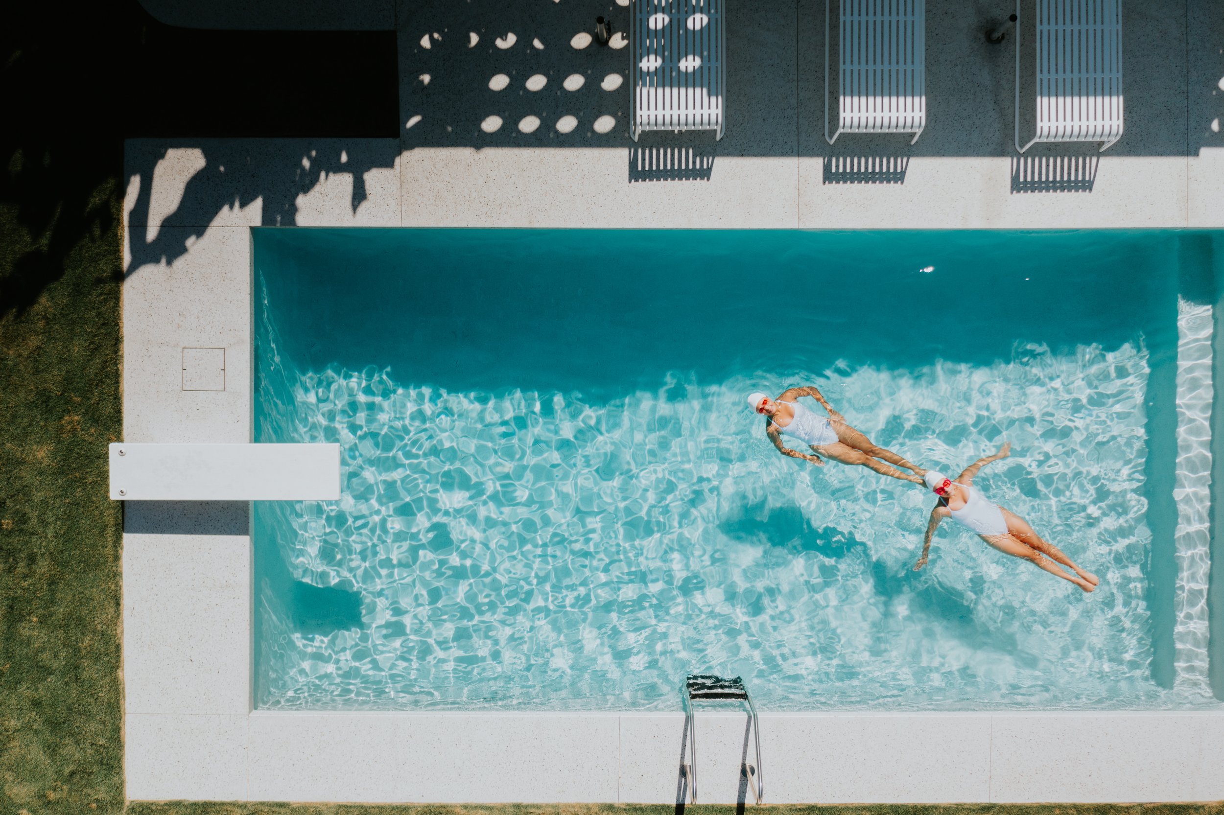 Aerial view of two people in white swimsuits floating in a swimming pool with a diving board and poolside chairs.