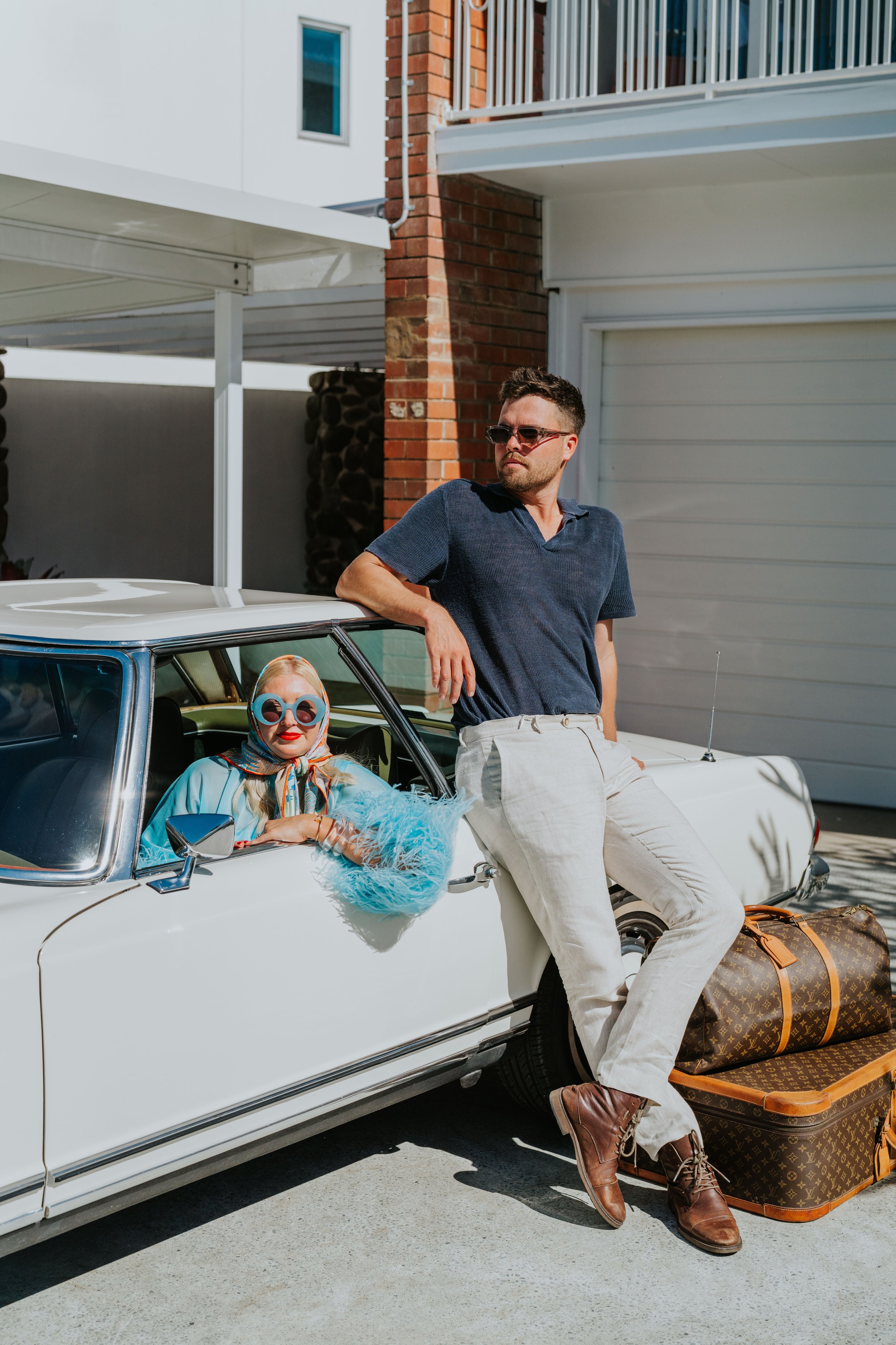 A woman wearing sunglasses, a headscarf, and a blue outfit sits in a vintage white car, while a man in sunglasses, a navy shirt, and light pants leans against the car. They are outdoors near a brick building with two large pieces of brown luggage on