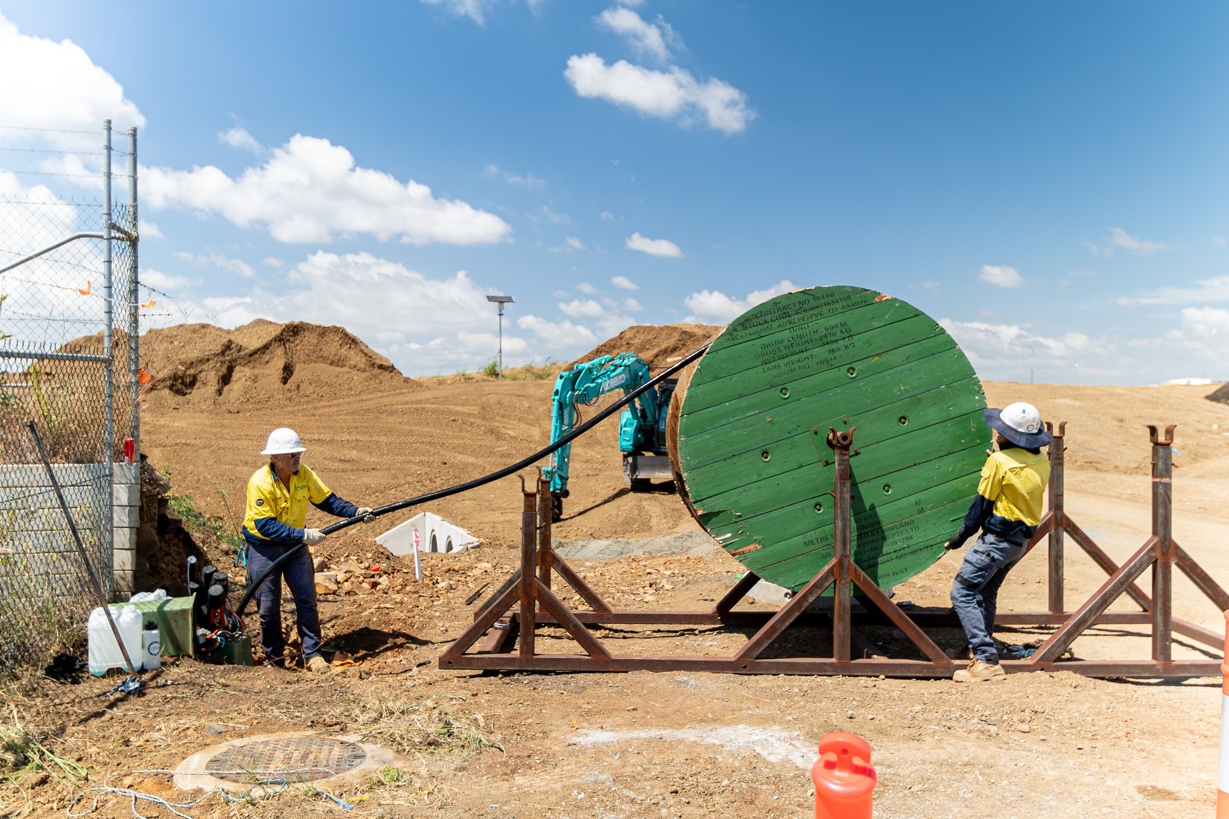 Two construction workers in yellow shirts and helmets pulling a large cable from a green spool at a construction site.