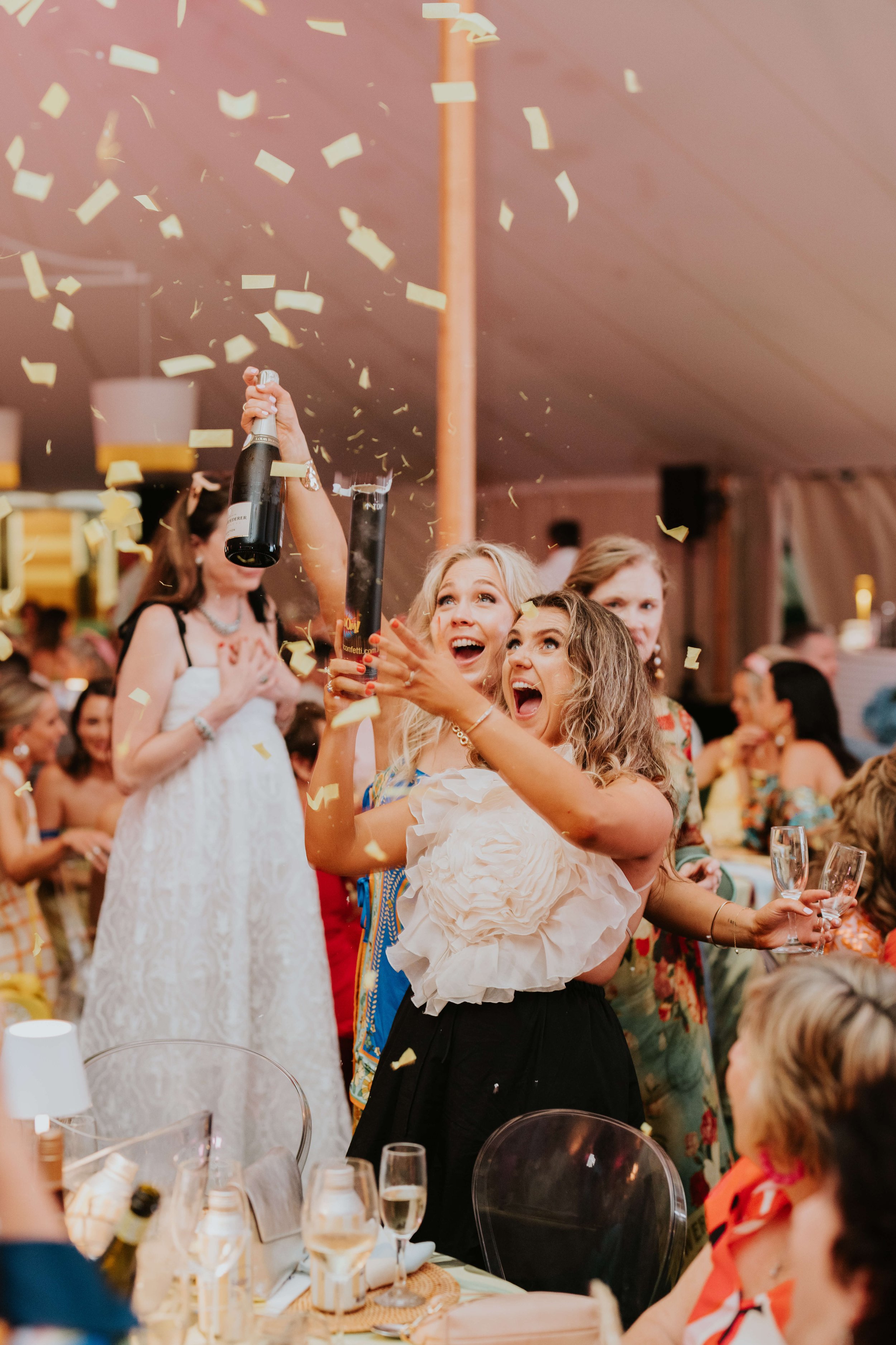 A lively celebration scene with women excitedly holding champagne bottles and glasses while yellow confetti falls around them, creating a festive atmosphere indoors.