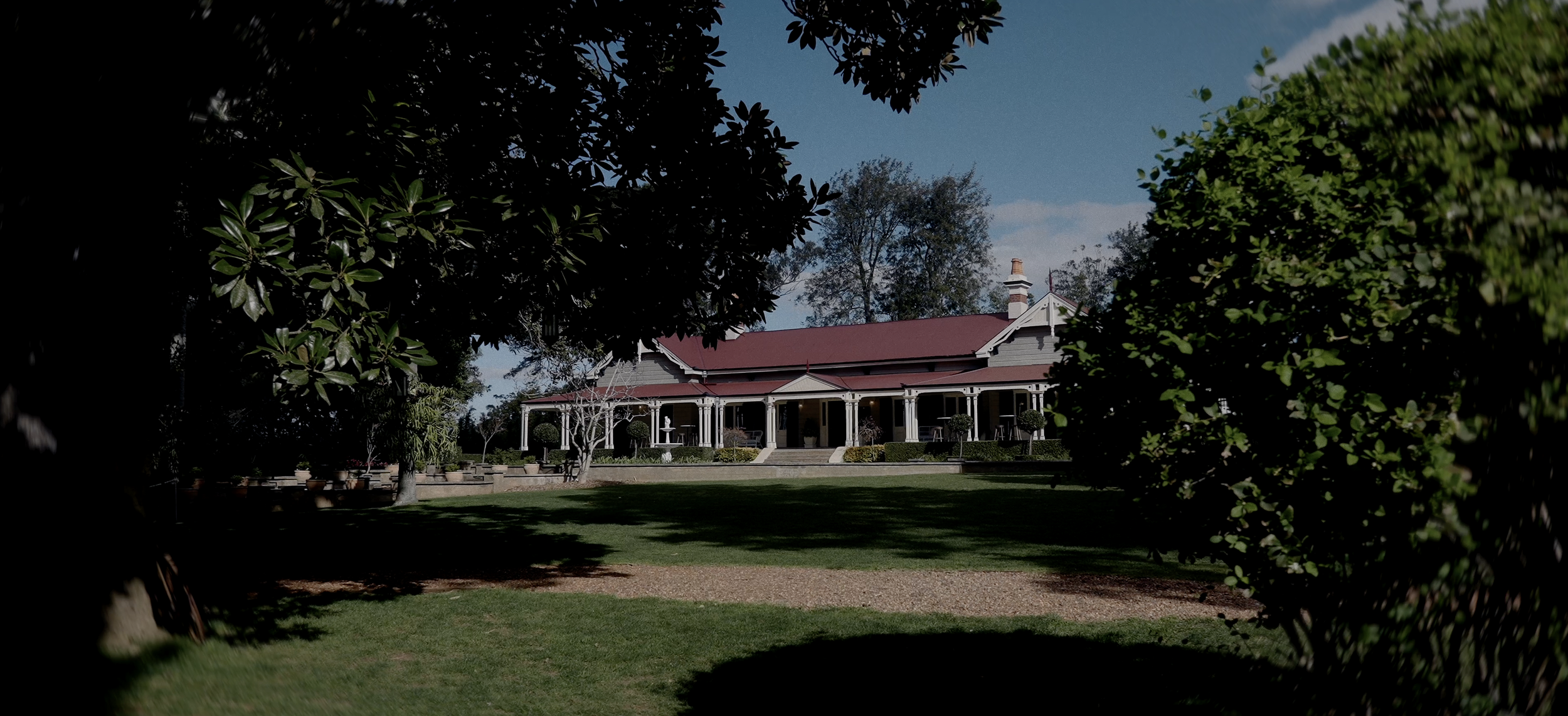 Historic house with red roof surrounded by trees and lawn, clear sky in background.