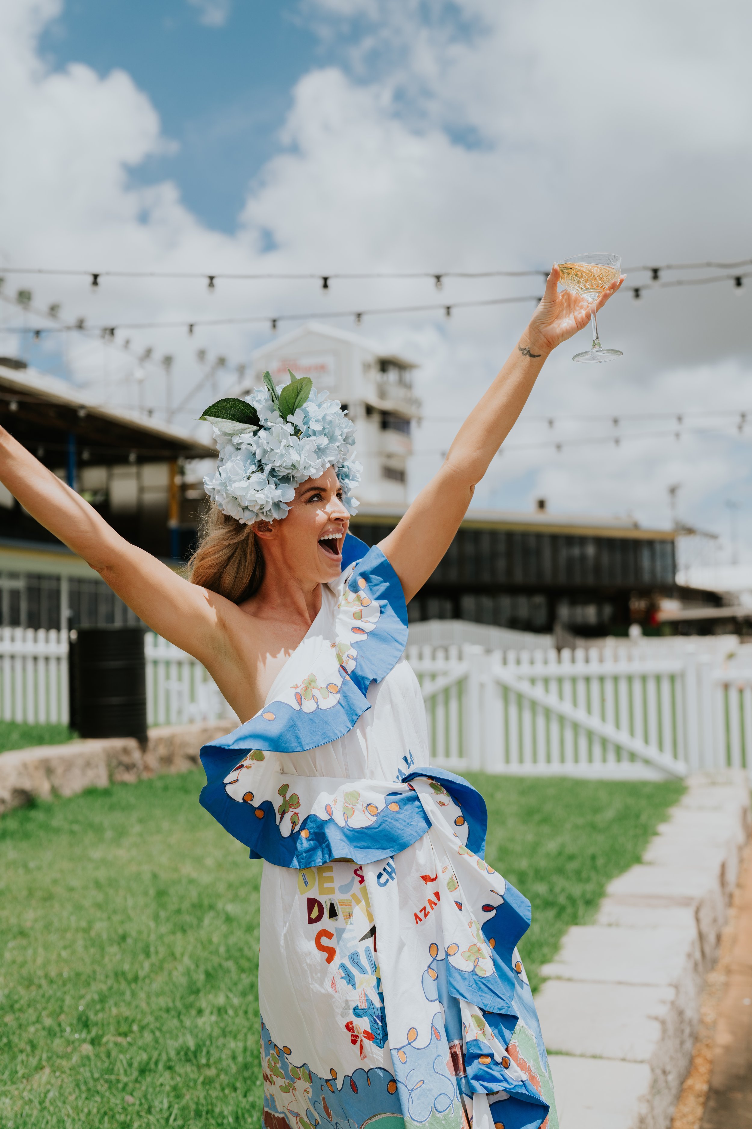 Woman celebrating outdoors, wearing a floral headpiece and colorful dress, holding a glass of champagne.