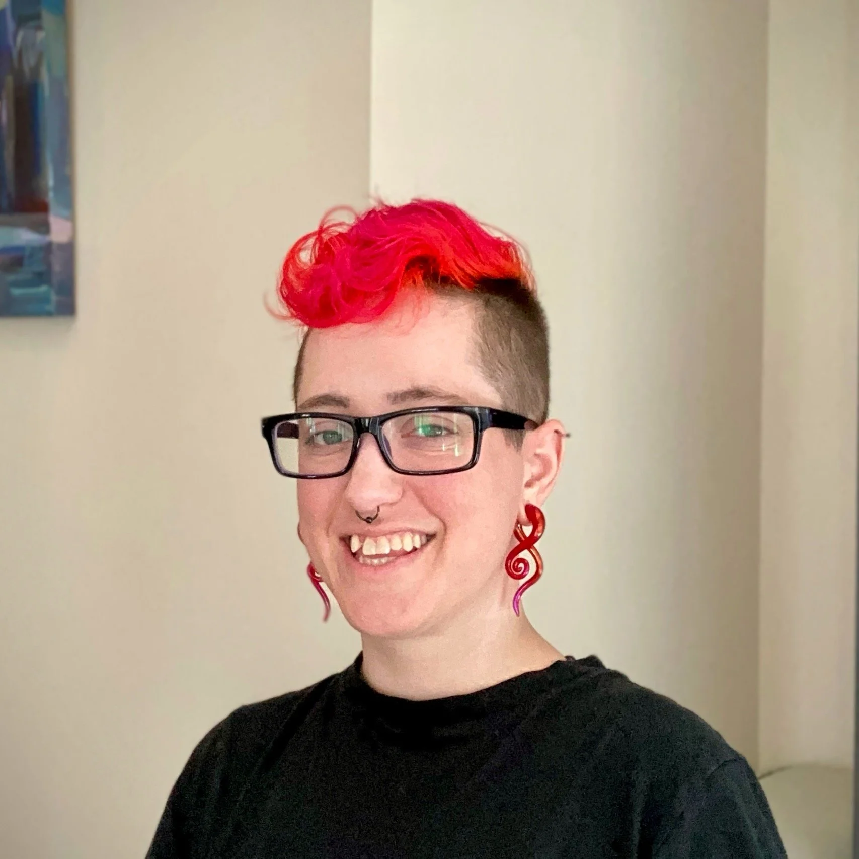 Person with short dark hair styled with bright red curly top, wearing black glasses, red swirl earrings, a nose septum ring, and a black shirt, smiling in front of a neutral wall.
