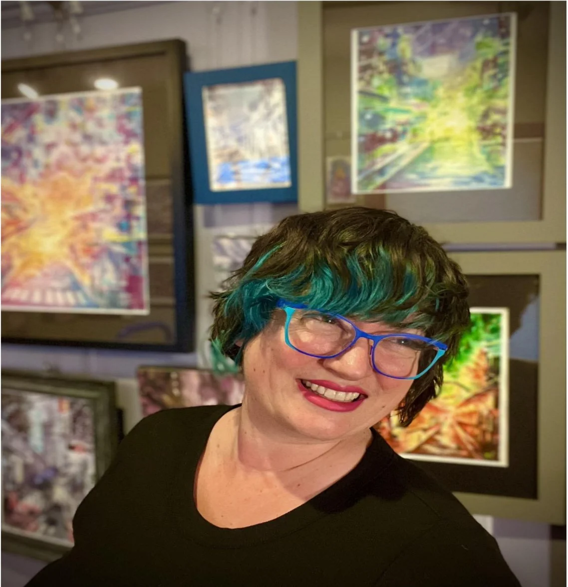 A woman with short dark hair with blue and green highlights, wearing blue glasses and a black shirt, smiling in front of colorful painting displays.
