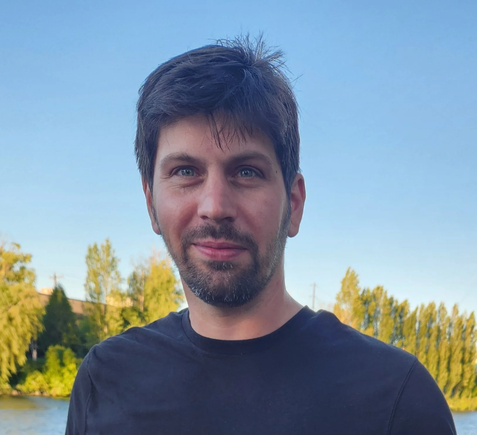 A man with dark hair, a beard, and a mustache, looking into the camera with a slight smile, outdoors near a body of water with trees in the background under a blue sky.