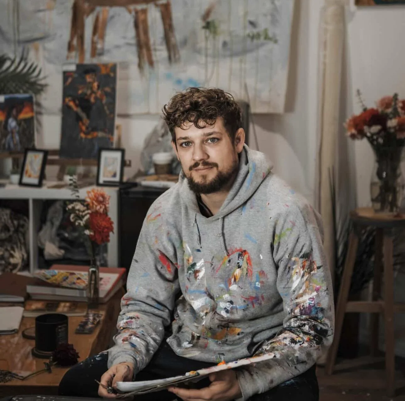 A man with curly hair and a beard sitting in his art studio, wearing a paint-splattered hoodie, surrounded by paintings, flowers, and art supplies.