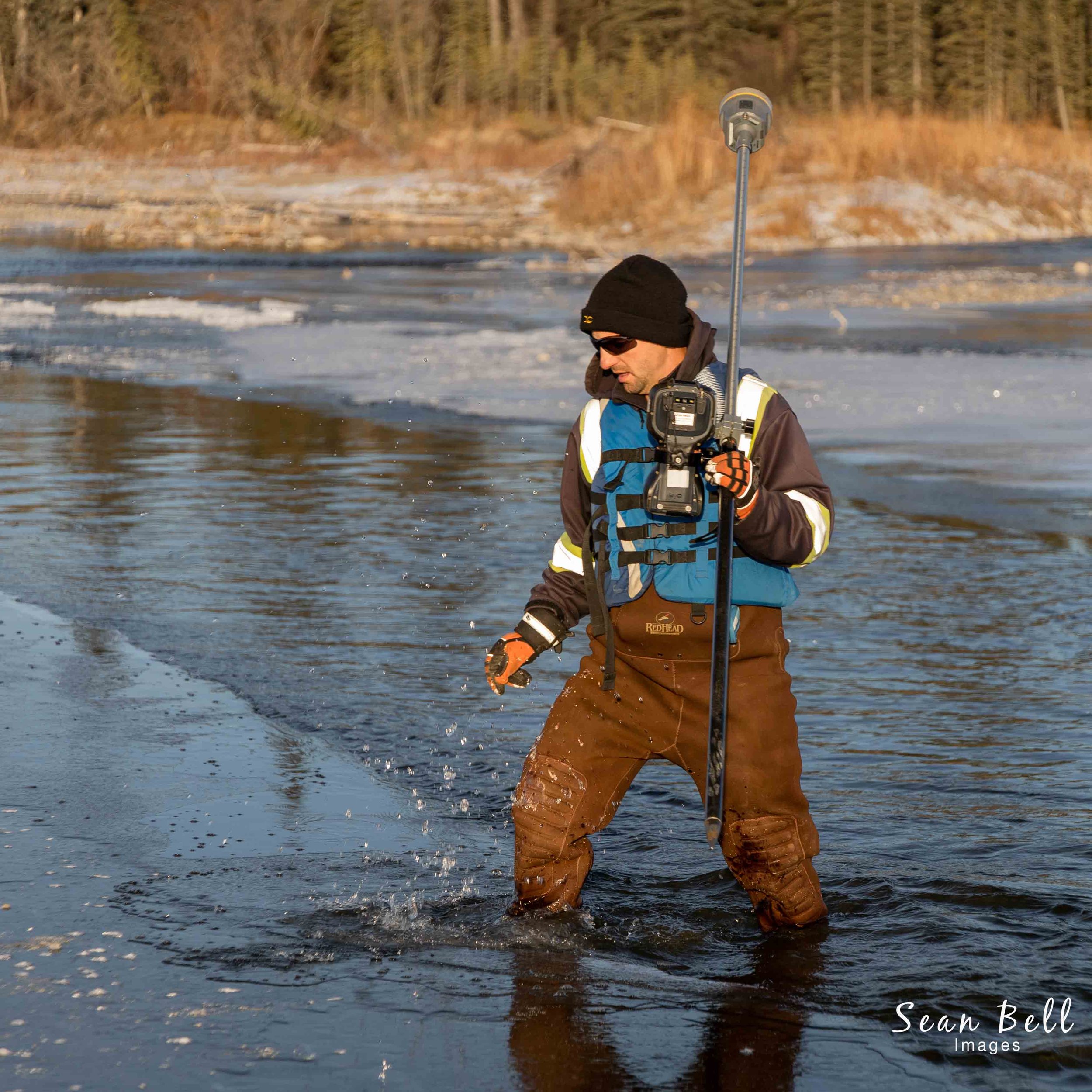 Action photo of a field professional in waders standing in a shallow, icy river, holding advanced surveying equipment. Highlights environmental fieldwork, waterway assessments, and land surveying in challenging conditions, promoting expertise in envi