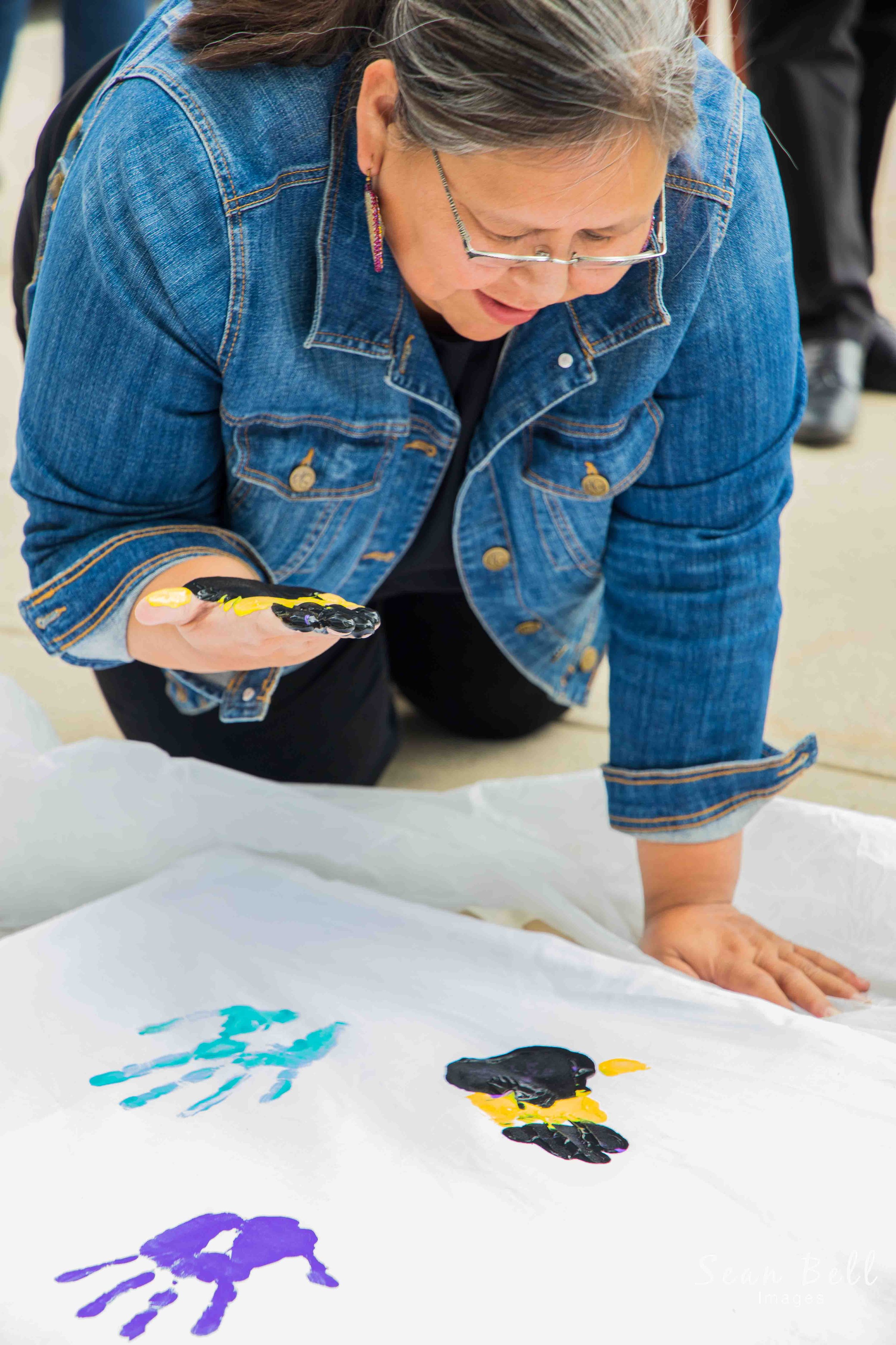 Engaging photo of a woman leaning over a painted surface, creating colourful handprints during a community art activity. Highlight creativity, cultural expression, and community engagement, promoting inclusive events, artistic initiatives, or diversi