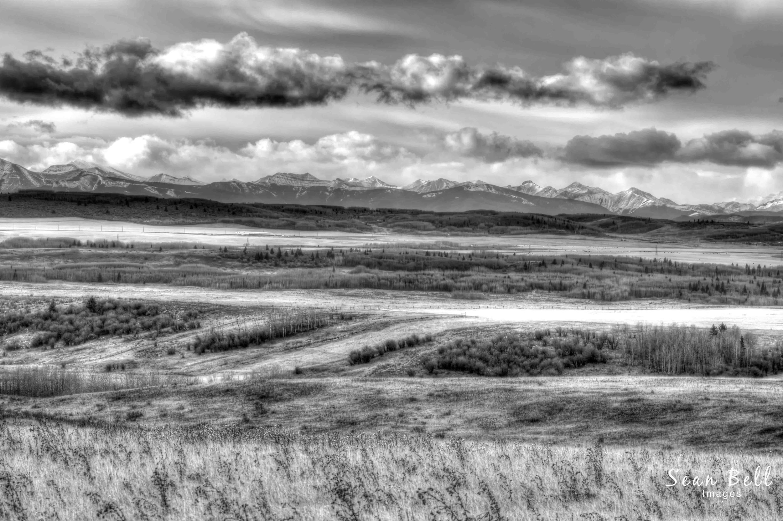 Black-and-white landscape photograph taken near Cochrane, Alberta, showcasing rolling fields leading to the dramatic silhouette of the Rocky Mountains under dynamic, textured clouds. This image captures the timeless beauty and contrast of Alberta's n