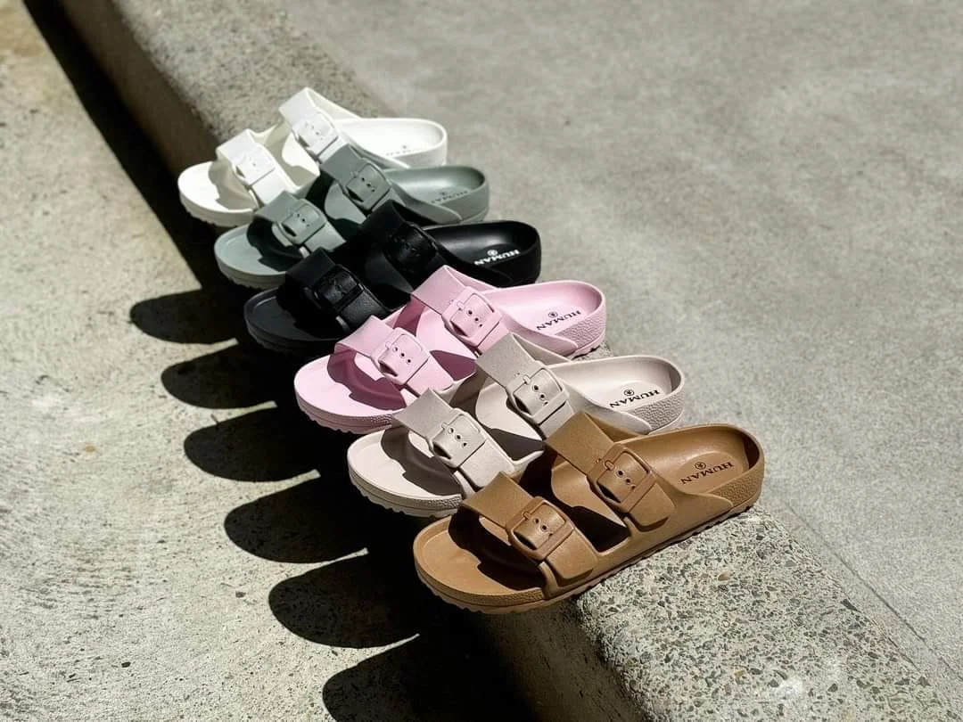 A row of colorful sandals in white, gray, black, pink, beige, and brown lined up on a concrete surface, with shadows cast by sunlight.