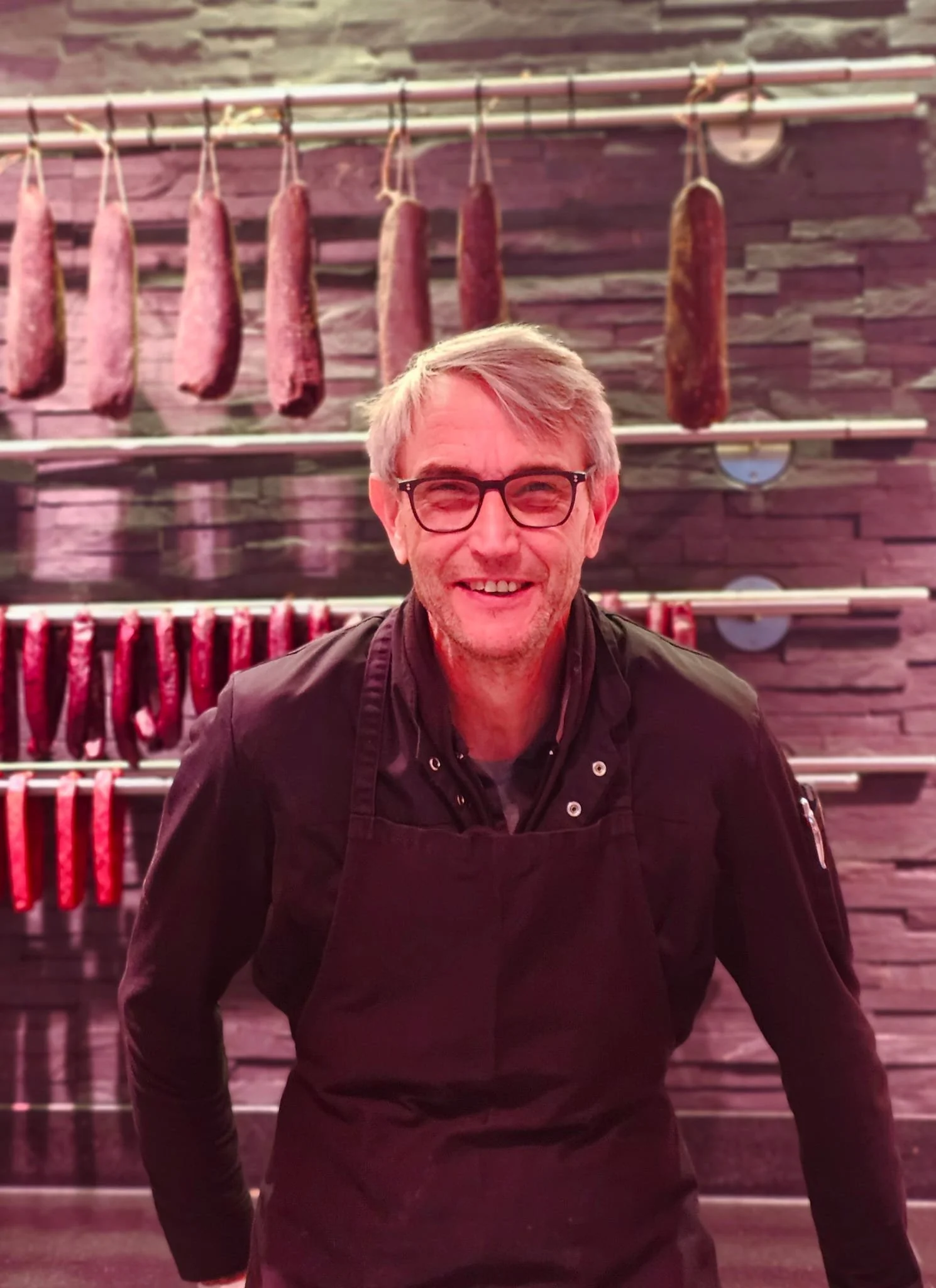 Lächelnder Mann in Schwarzkochjacke und Brille vor einer Wand mit hängendem Fleisch