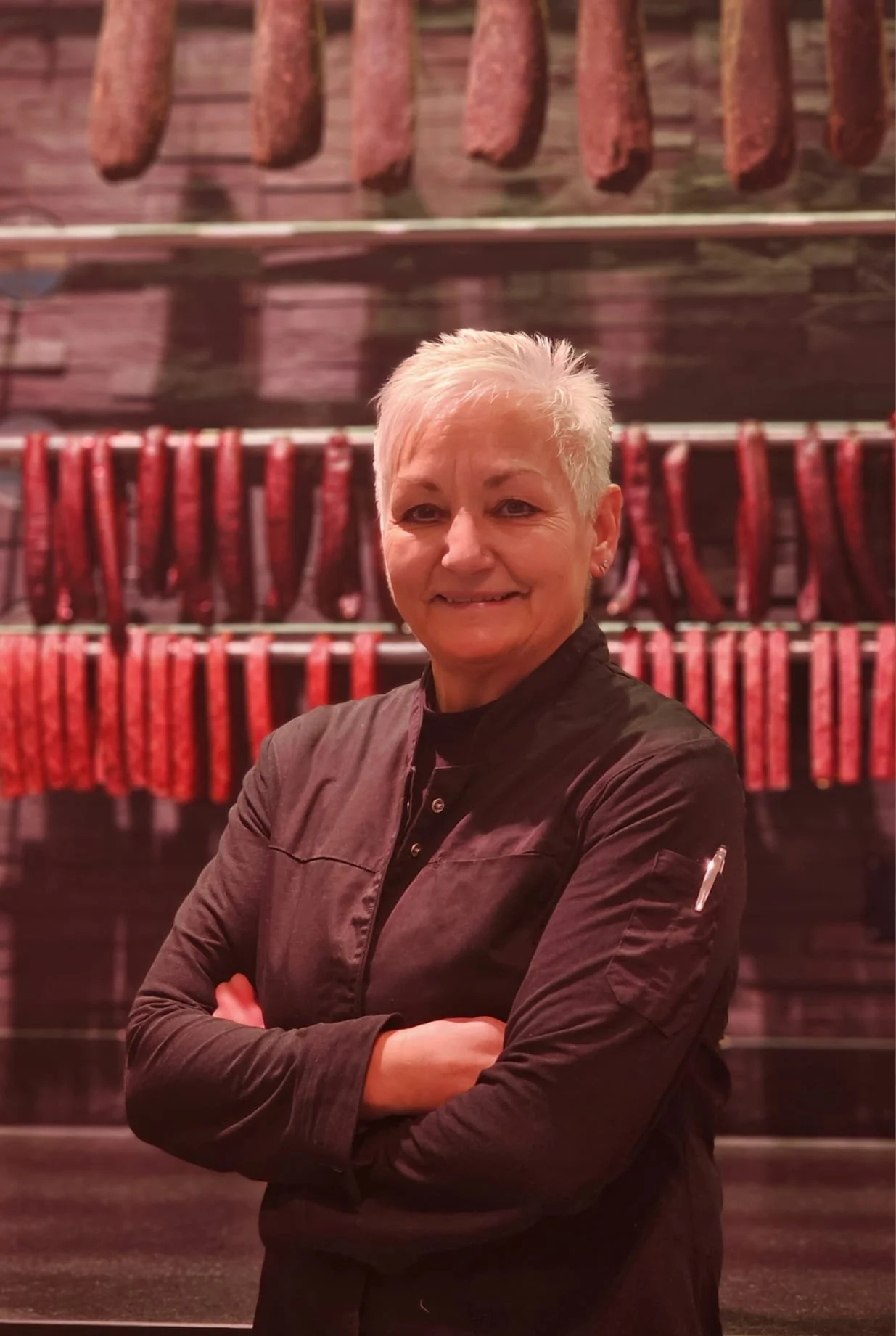 Eine ältere Frau mit kurzen, silbernen Haaren, trägt eine schwarze Kochjacke, steht vor einer Wand mit aufgehängten Wurstwaren in einem Metzgerladen oder einer Fleischerei, lächelt und hat die Arme verschränkt.