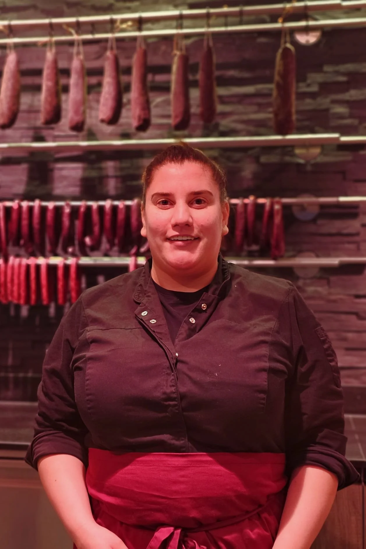 Eine Köchin steht vor einer Wand mit aufgehängtem rohem Fleisch, darunter getrocknetes Fleisch.