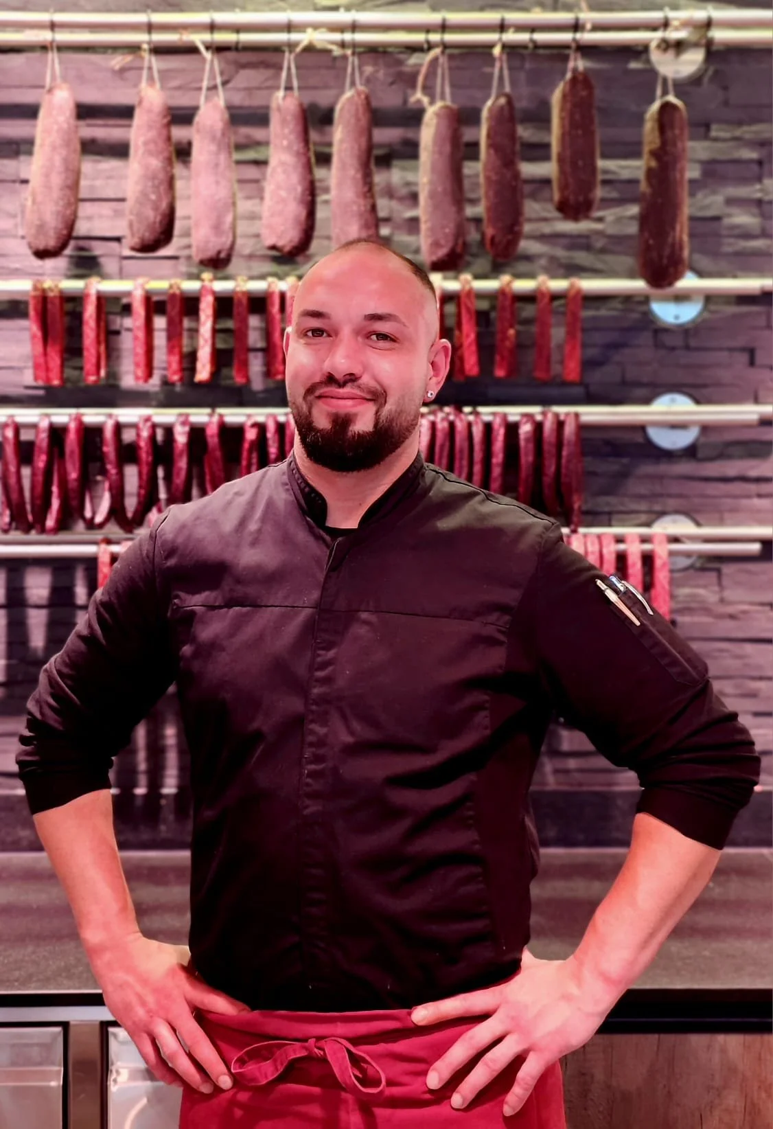 Ein Küchenchef steht vor einer Wand mit verschiedenen Wurstsorten, die an Haken hängen.