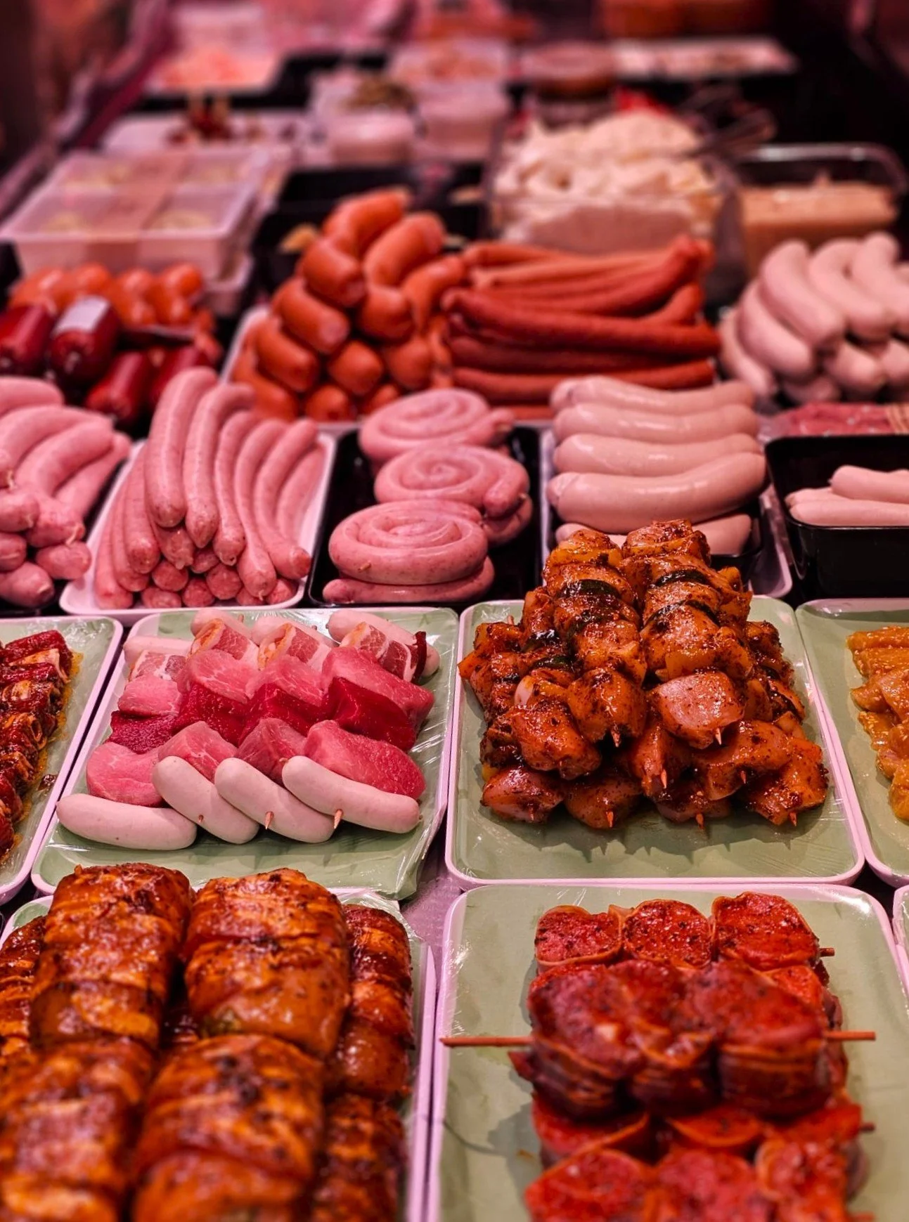 Verschiedene Fleisch- und Wurstwaren auf Märkten, darunter Bratwürste, Hühnerfleisch, gefüllte Fleischspieße, Hähnchenschenkel, und andere Fleischprodukte auf Tellern und in Behältern.