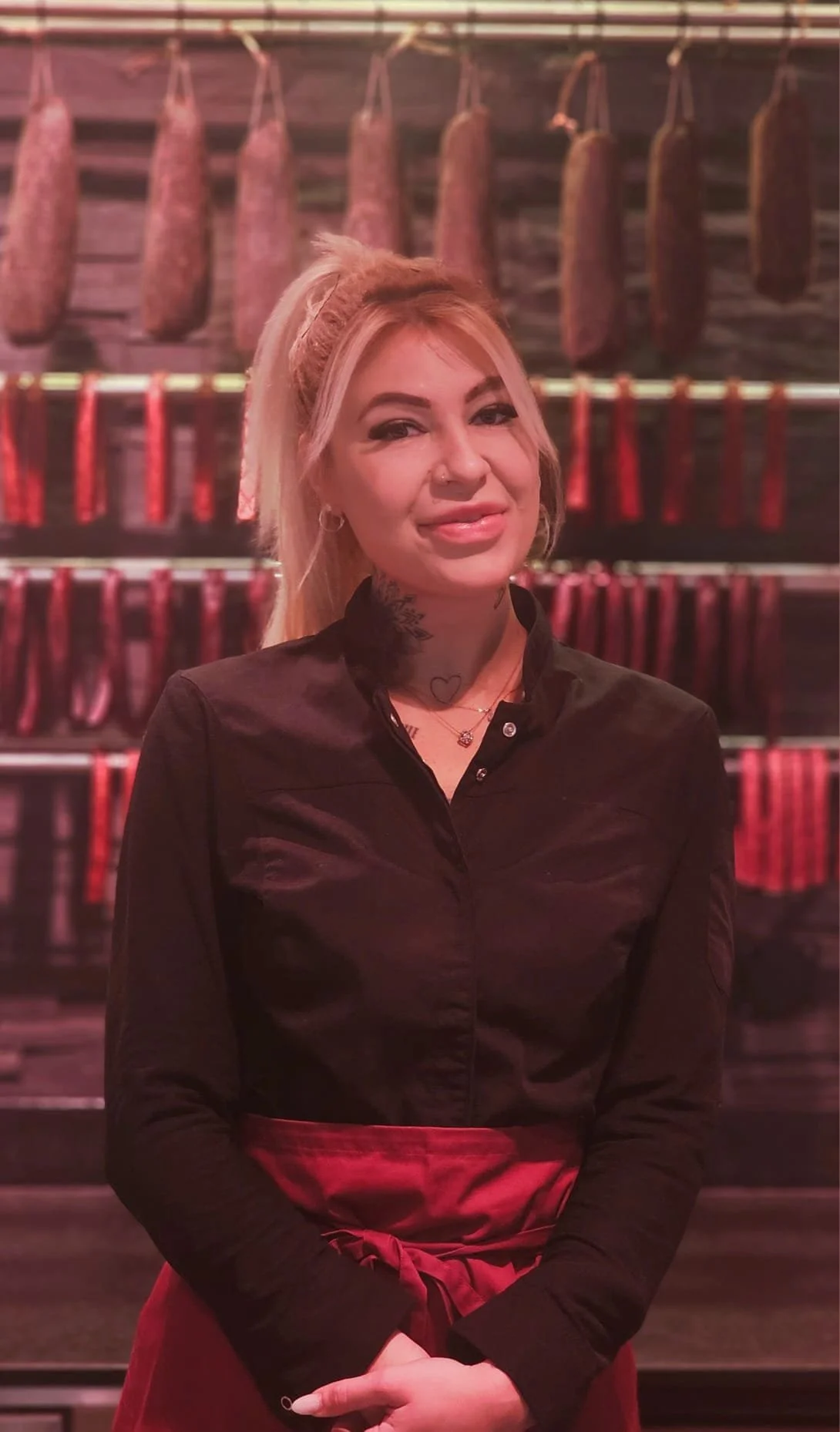 Eine Frau steht vor einer Wand mit aufgehängtem Fleisch, vermutlich Wurst oder Schinken, in einem Metzgerei-Shop.