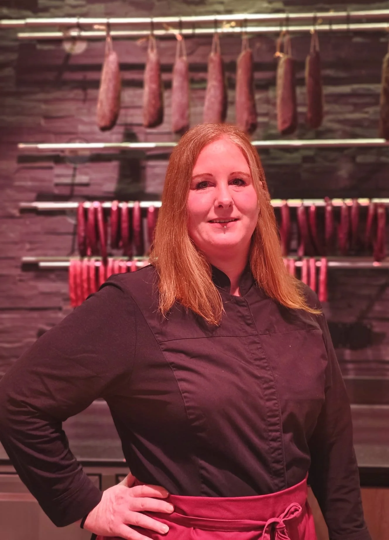 Eine Frau mit roten Haaren trägt eine schwarze Kochjacke und eine rosa Schürze. Im Hintergrund hängen verschiedene Wurstsorten an Haken an einer Wand in einer Metzgerei.