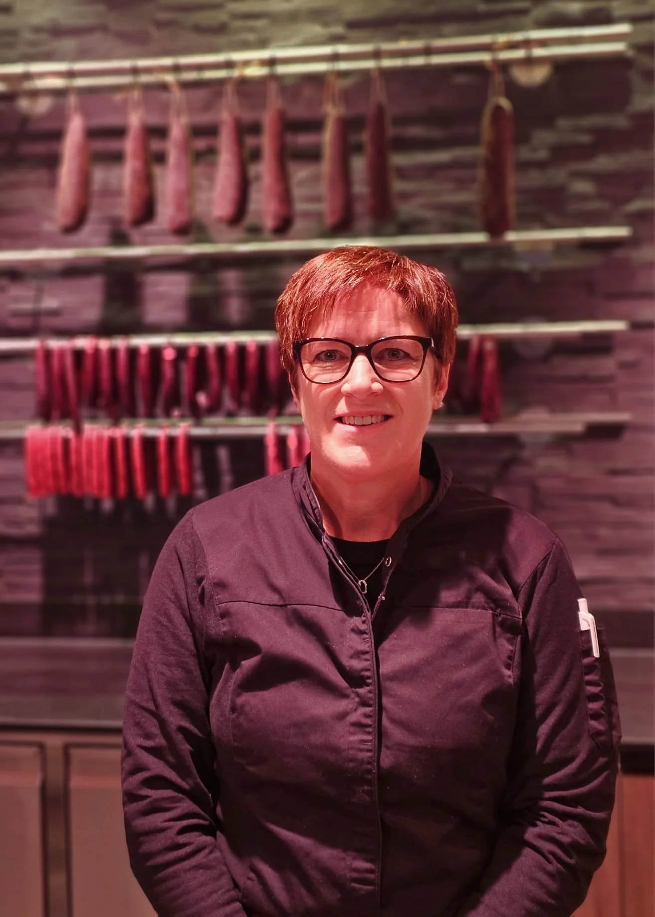 Eine Frau mit Brille und dunkler Jacke steht vor einer Wand mit verschiedenen Wurstsorten, die auf Holzbrettern hängen.