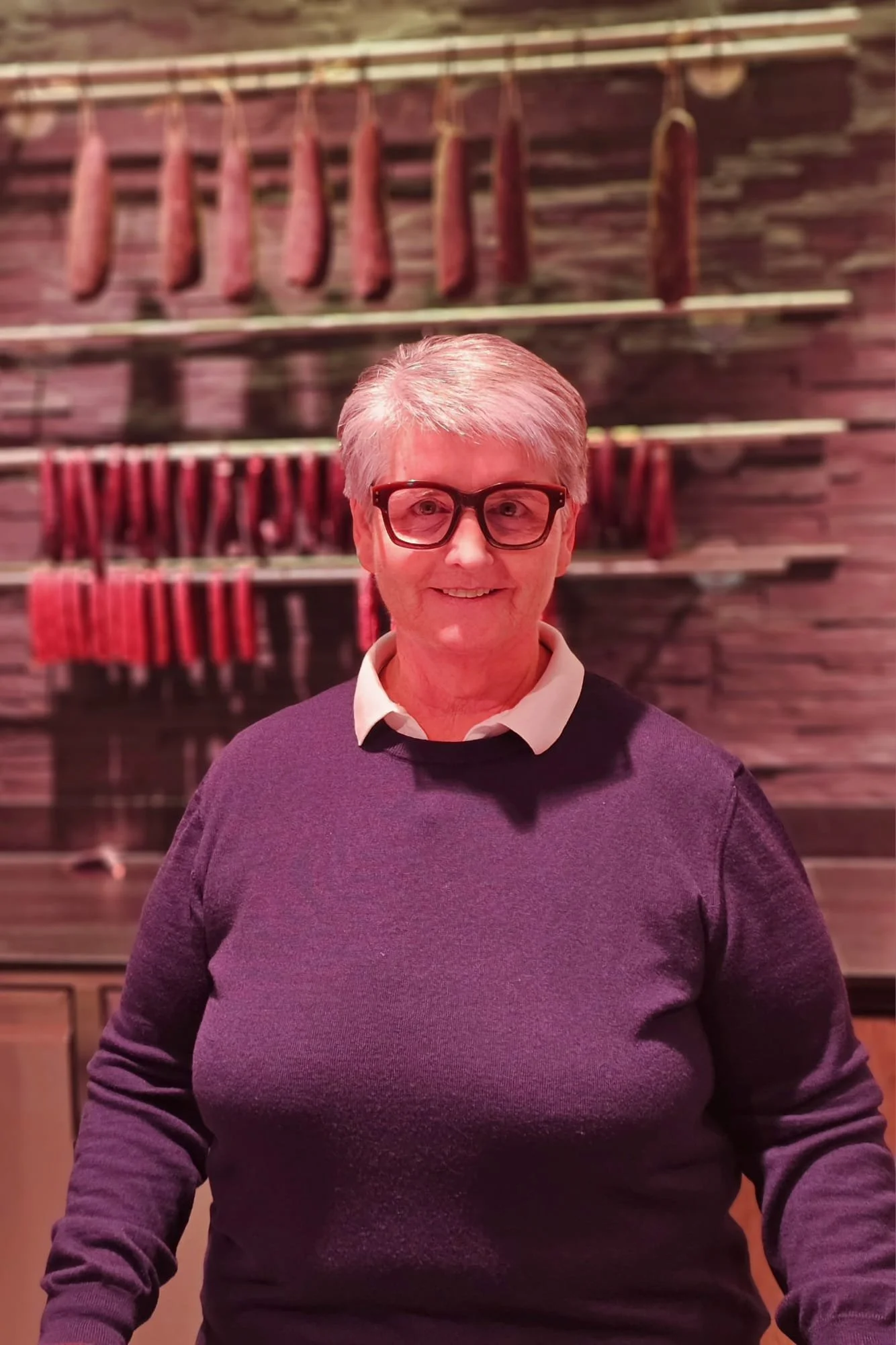 Eine lachende ältere Frau mit kurzen grauen Haaren, Brille und einem dunkelvioletten Pullover steht vor hängenden Wurstsorten an einer Holzwand.
