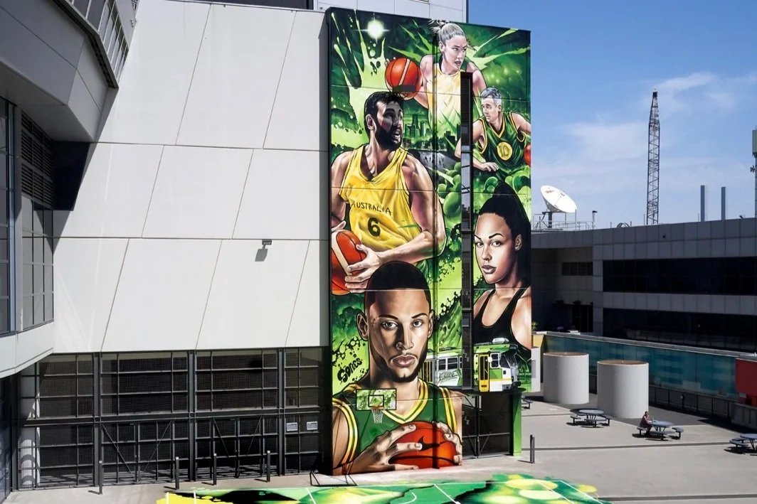 Colorful mural on a building depicting several basketball players, including a man in a yellow jersey, a woman with short curly hair, and another woman with long hair, with green background art. The mural also includes a basketball and a bus below it in an outdoor urban setting.