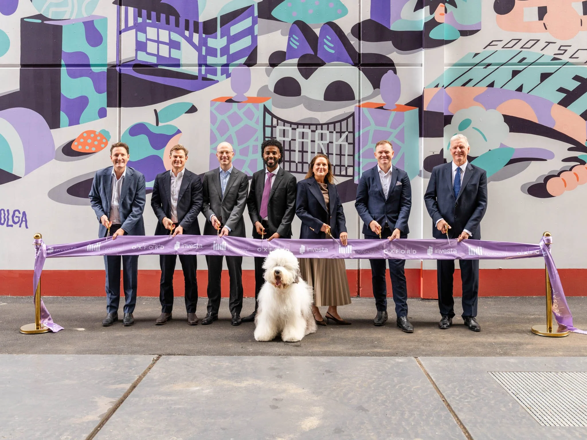 A group of seven people and a large white dog standing behind a purple ribbon at a ribbon-cutting event in front of a colorful mural.