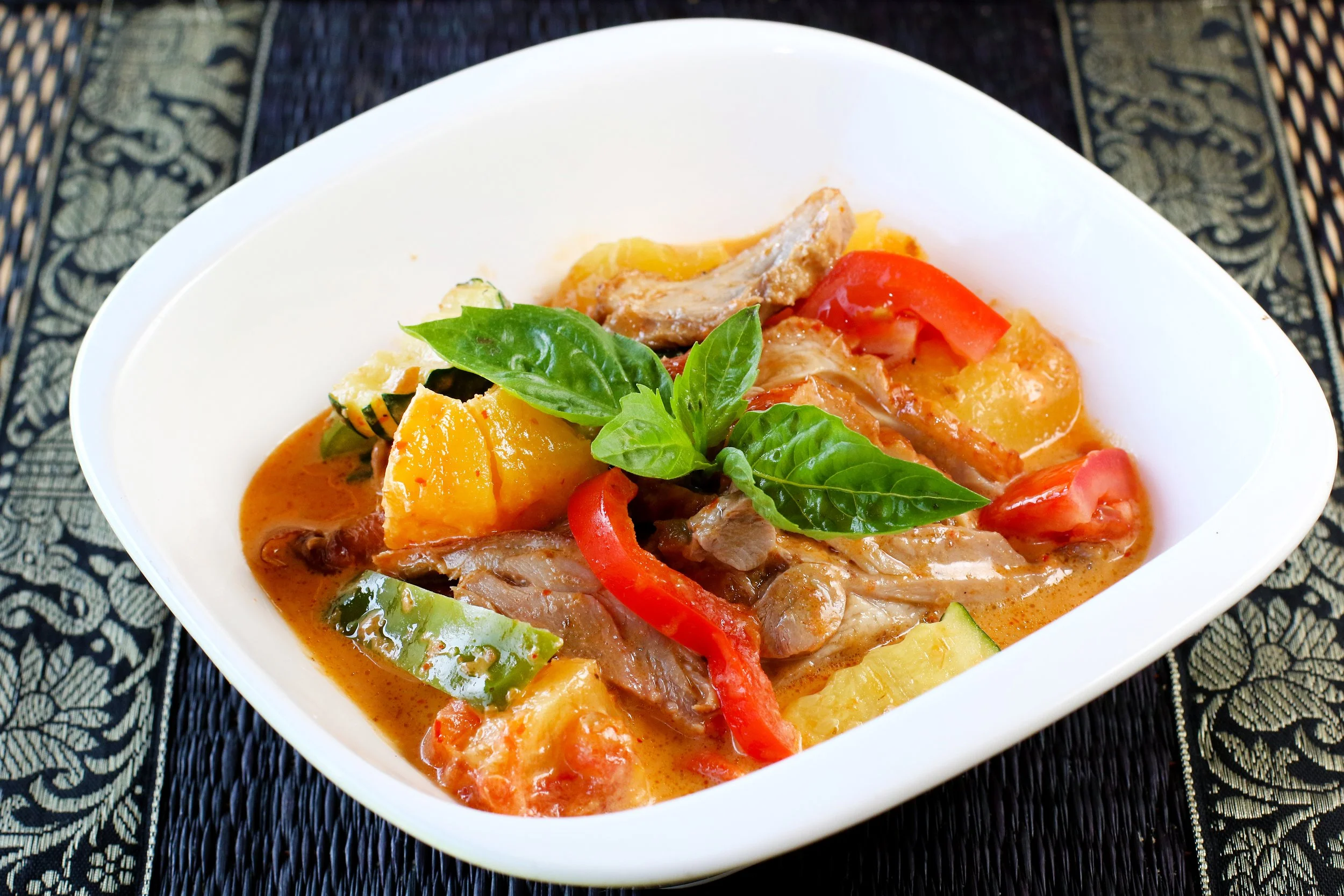 A bowl of roasted duck curry with red and green peppers, potatoes, zucchini, and basil leaves, served on a decorative table