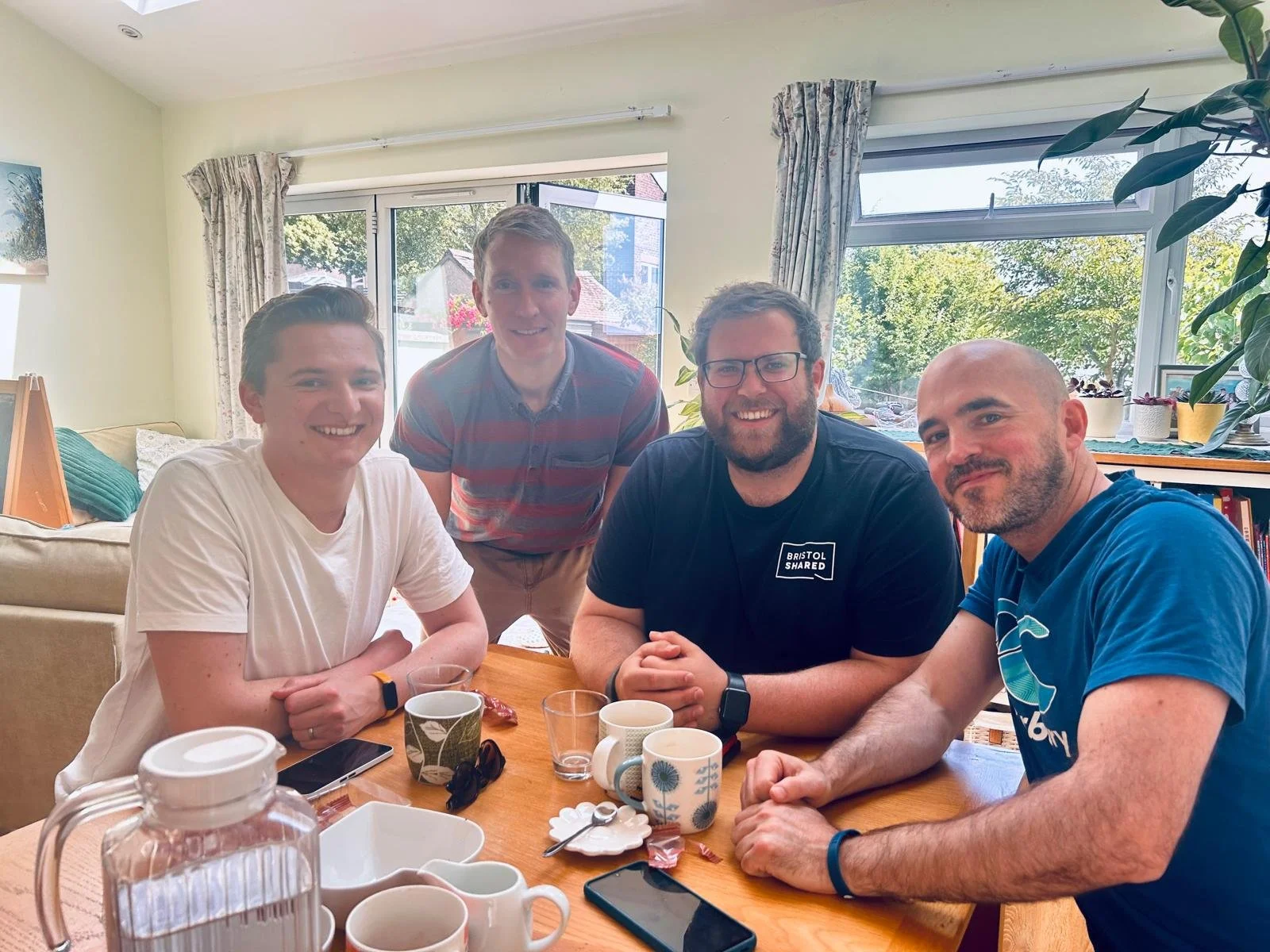 Four men sitting and standing around a dining table with mugs, a glass, and a pitcher, in a brightly lit room with large windows and curtains.