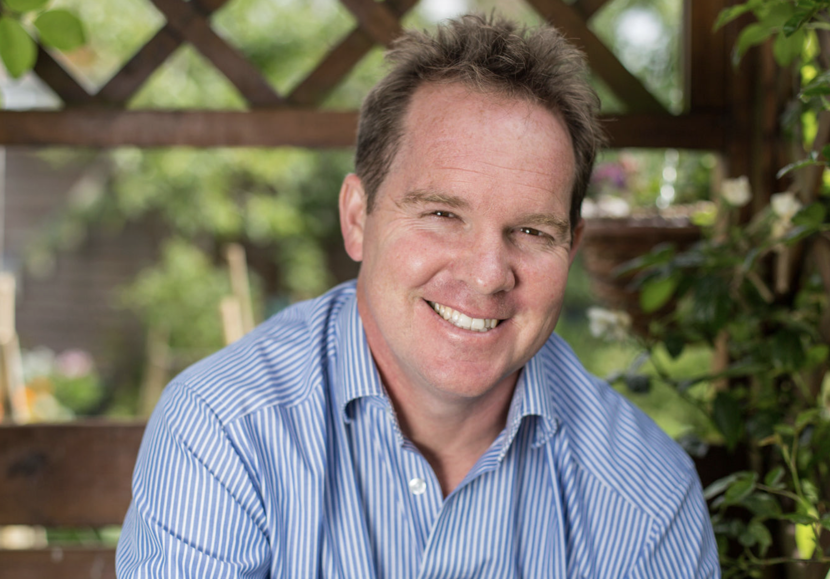 A smiling man with short brown hair, wearing a blue and white striped shirt, outdoors in a garden with trees and wooden fencing