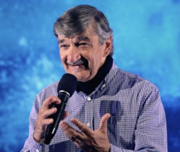 A man with gray hair and a mustache holding a microphone, speaking or performing on stage with a blue background.