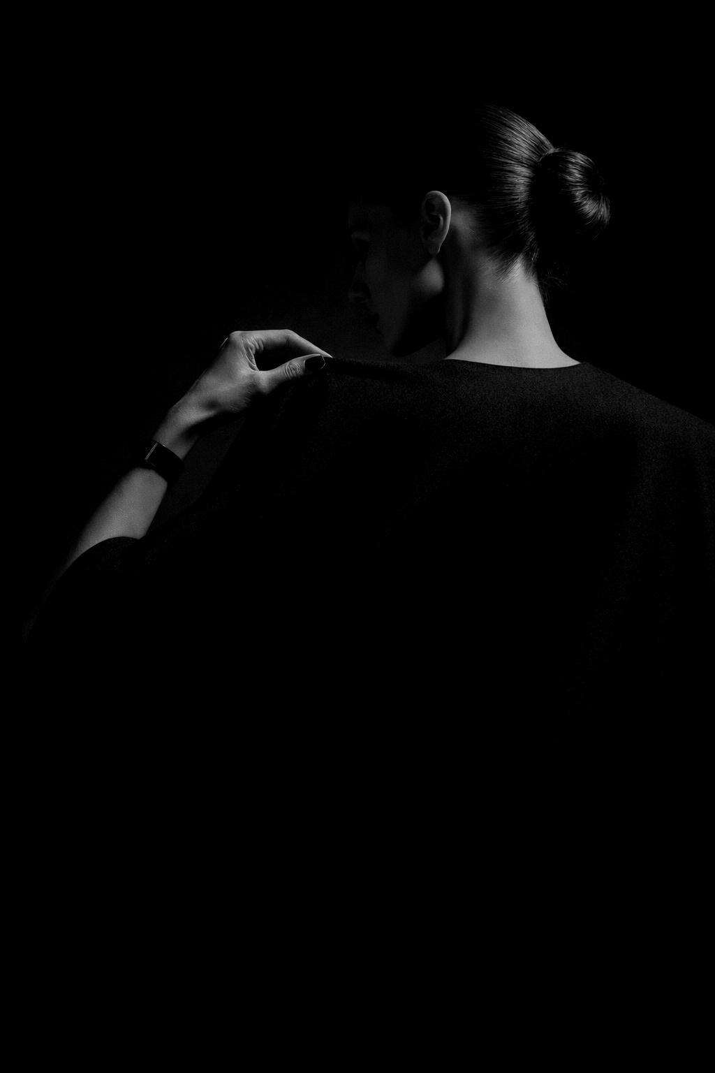 Back view of black cape on model in low lighting, with hand adjusting the shoulder in a minimalist fashion portrait
